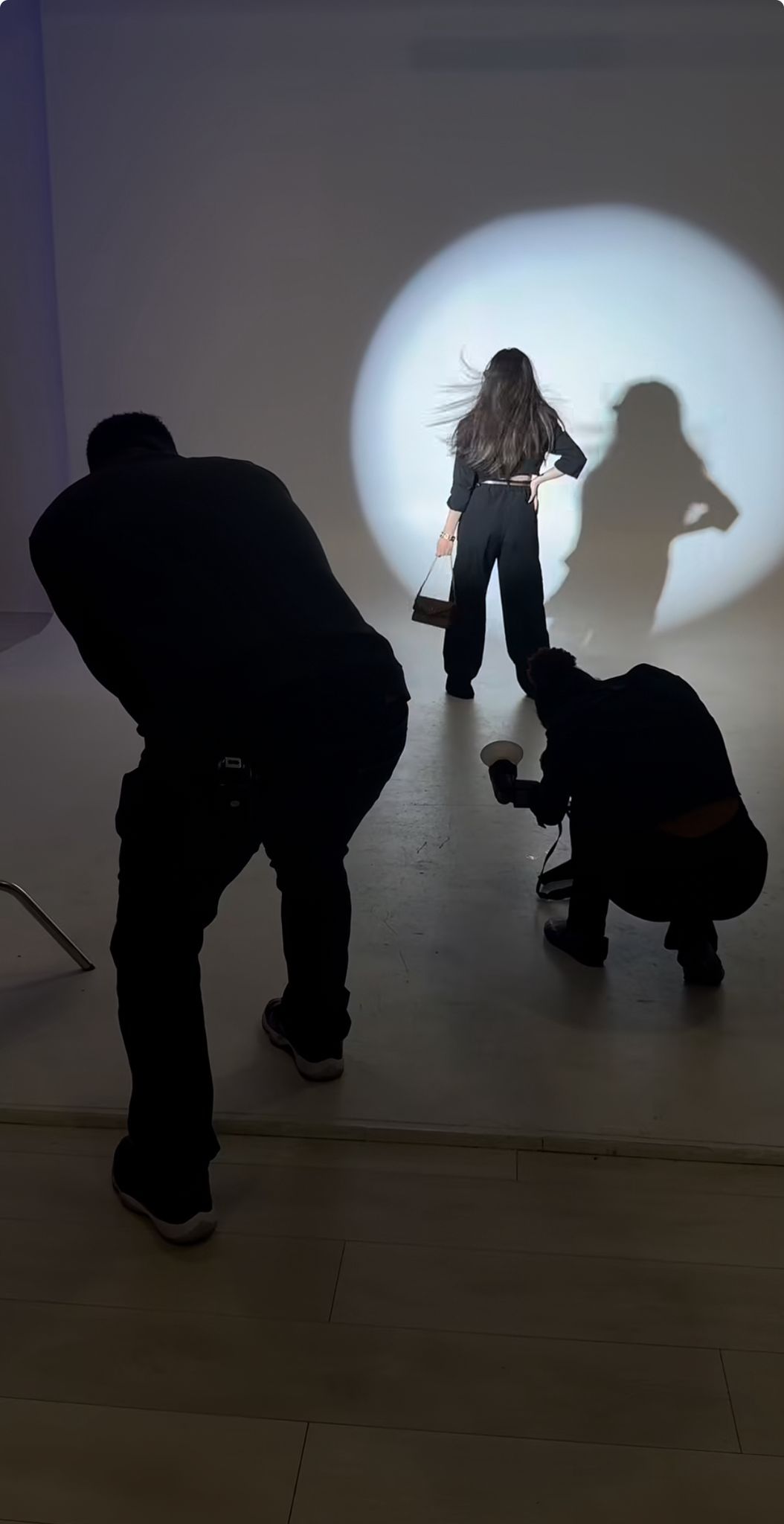 Silhouetted photographers capture a woman posing dramatically against a bright circular light projection on a white wall.