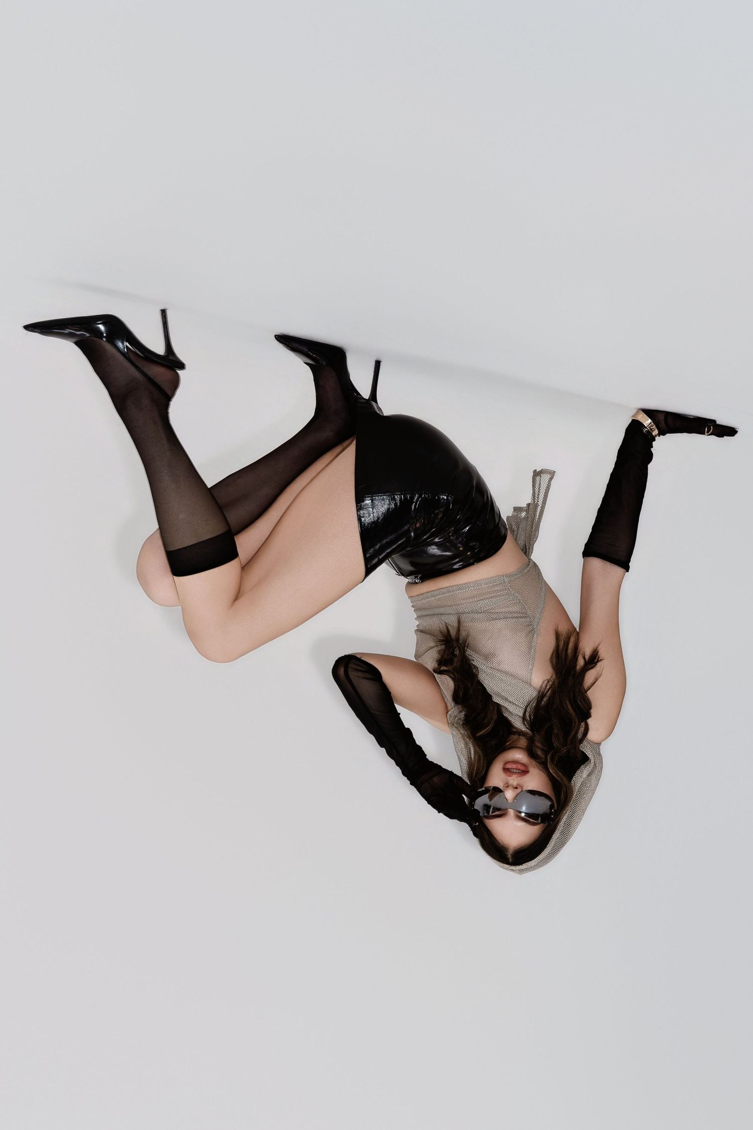 Woman in black latex outfit and heels posing upside down on white studio floor.