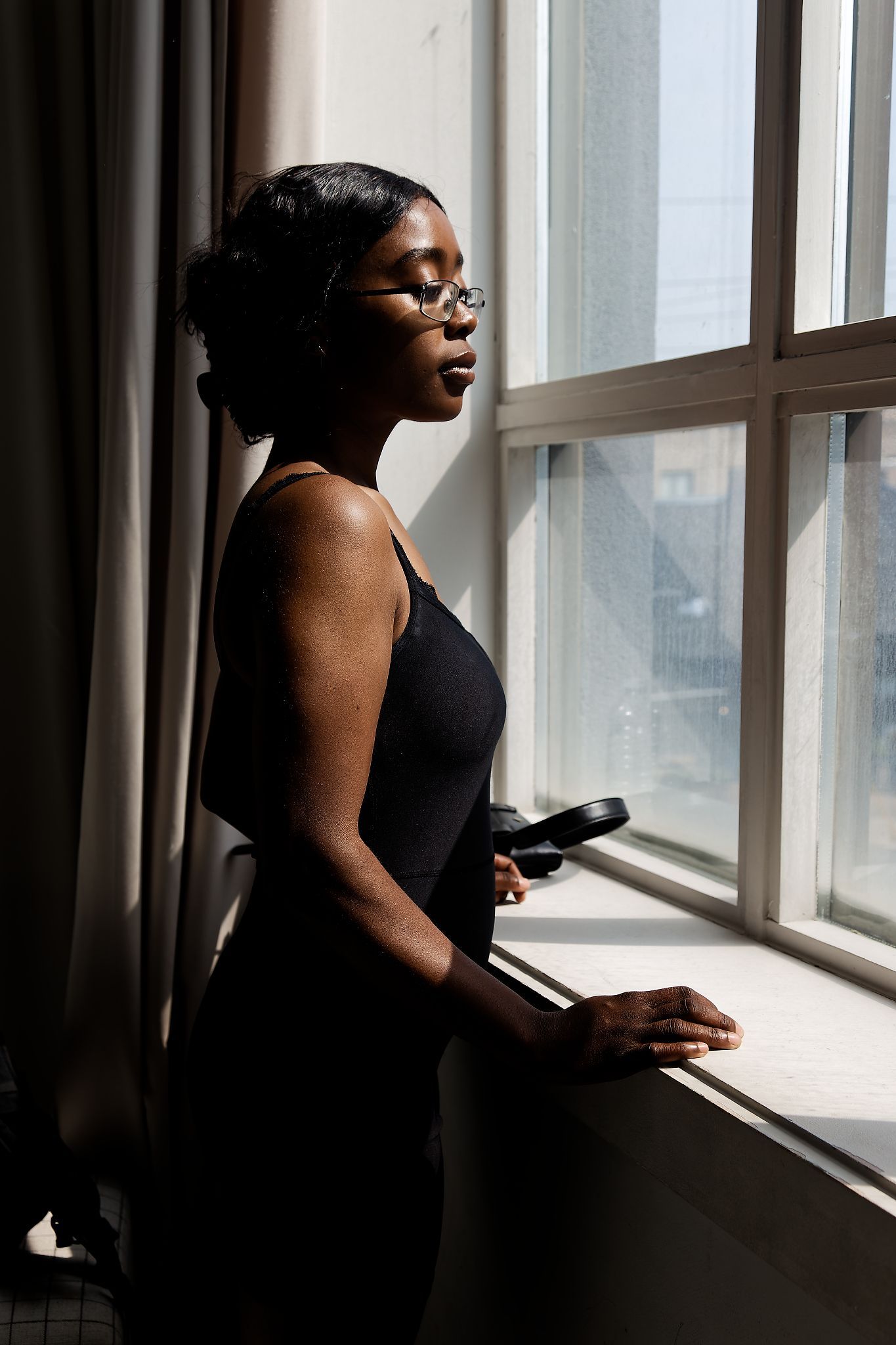 A Black woman in a black dress stands by a sunlit window, wearing glasses, holding a book in a contemplative pose.