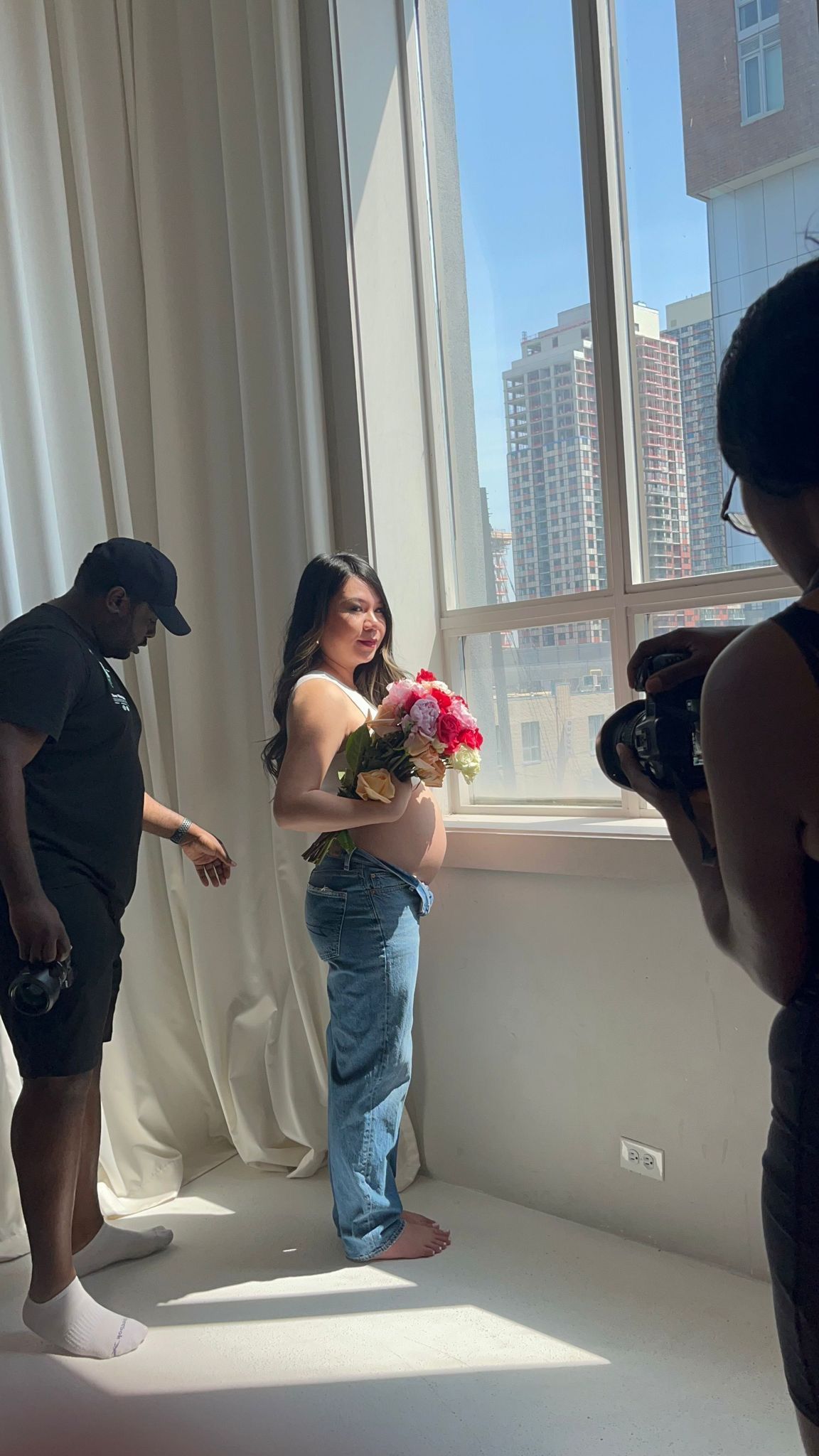 A pregnant woman holds a bouquet of flowers near a sunny window while photographers capture her maternity shoot.