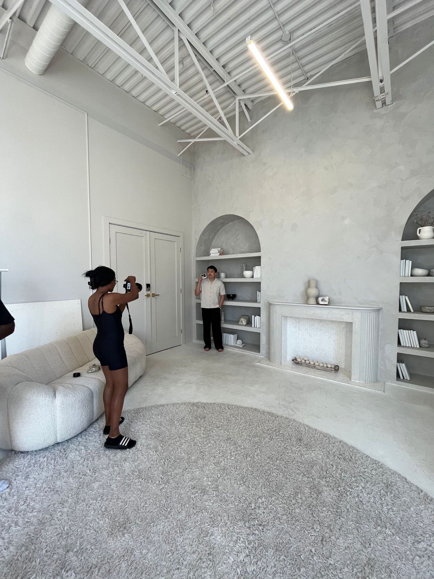 A photographer takes photos of a woman posing in a bright, modern white studio with arched shelving decor.