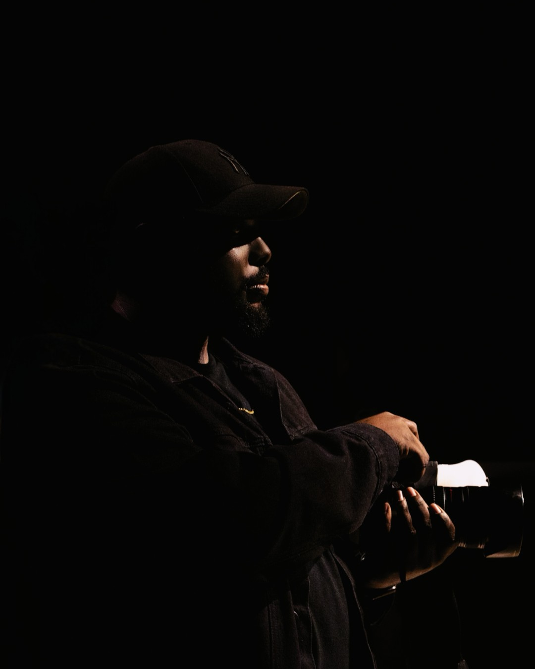 A silhouetted man wearing a cap sits with arms crossed in dramatic low-key lighting against a dark background.