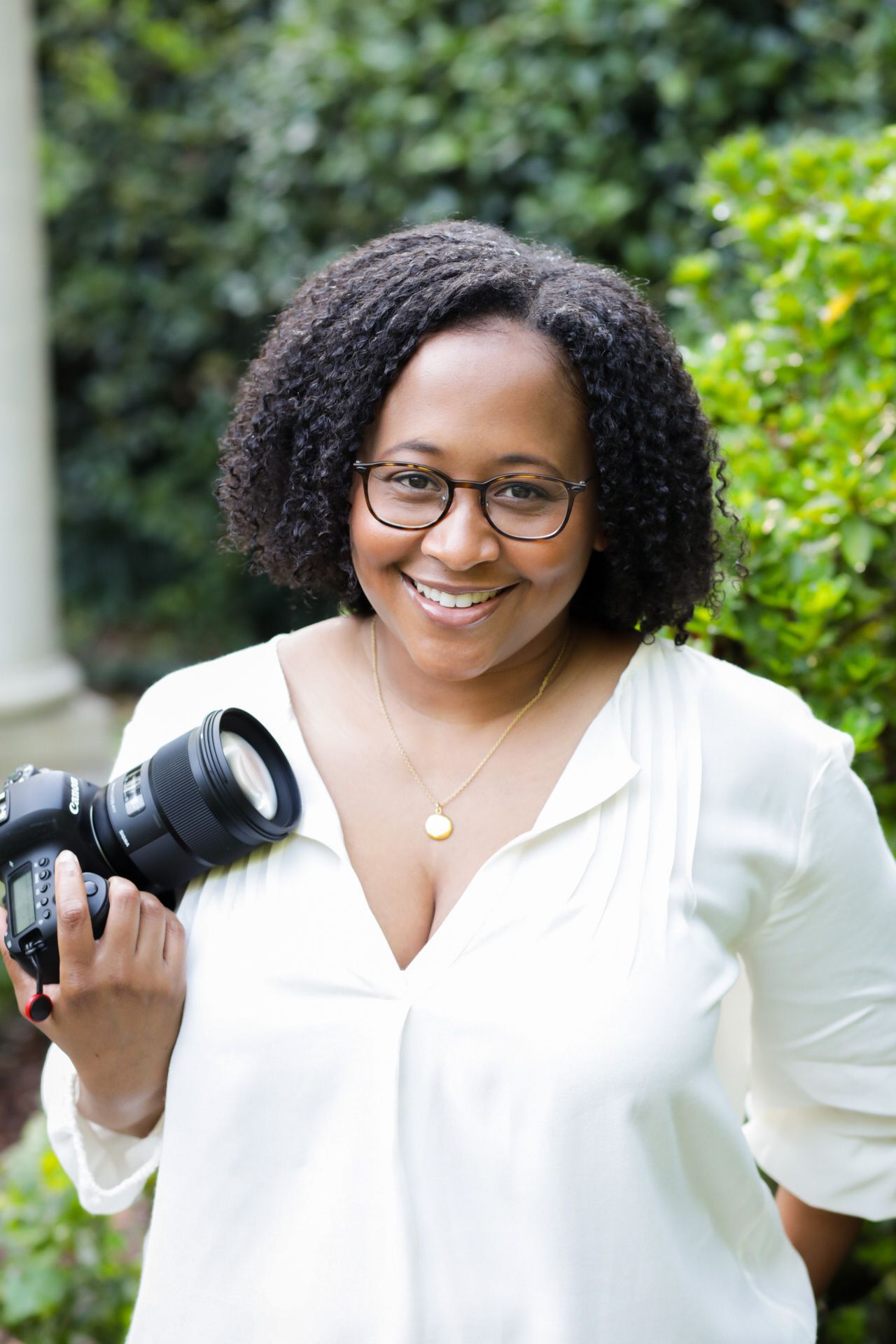 Professional photographer in white blouse with glasses holds camera while smiling in outdoor garden setting.