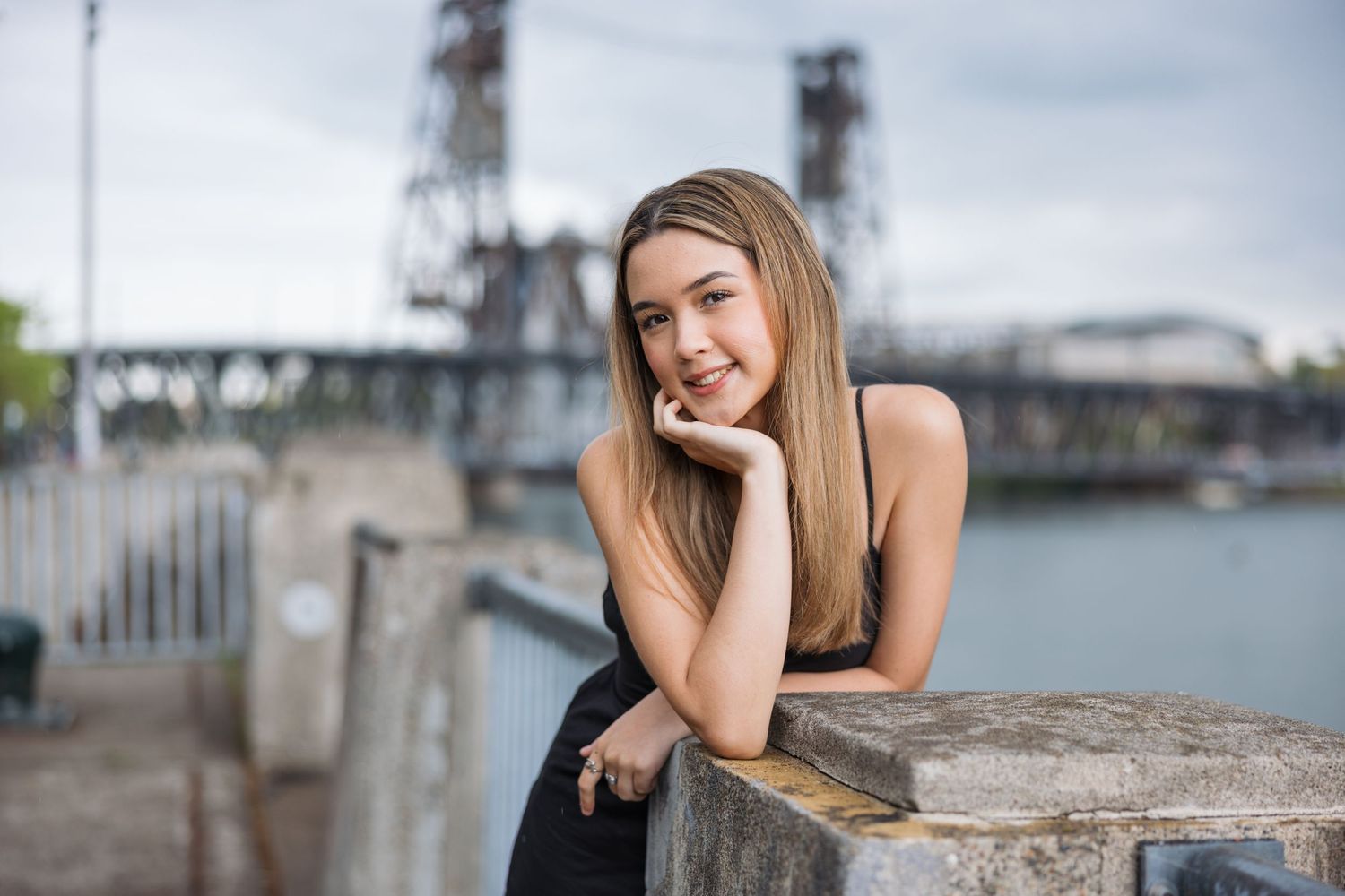 Woman in black leaning against stone railing with industrial bridge in background.