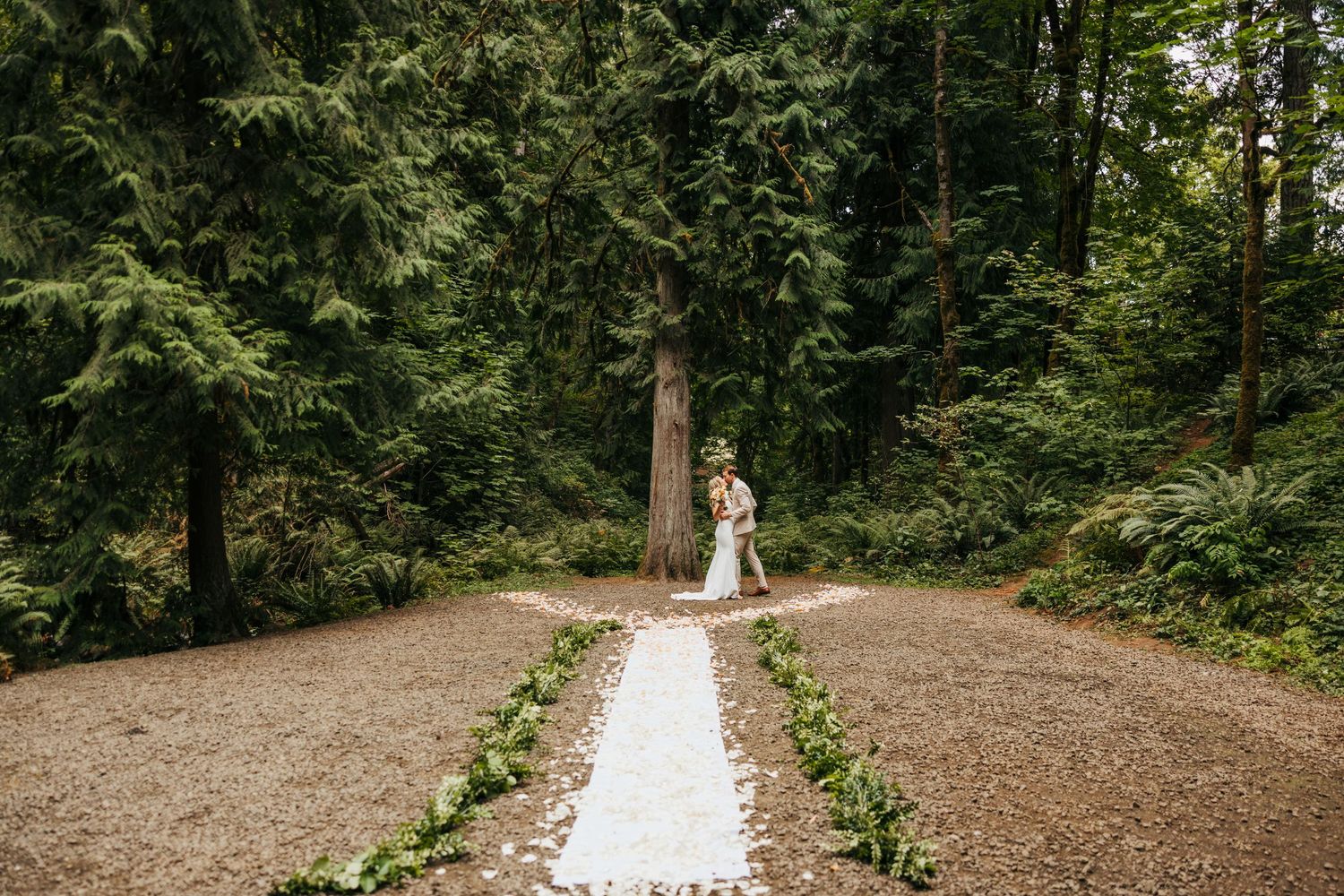 A white aisle runner lined with greenery leads through a forest setting.