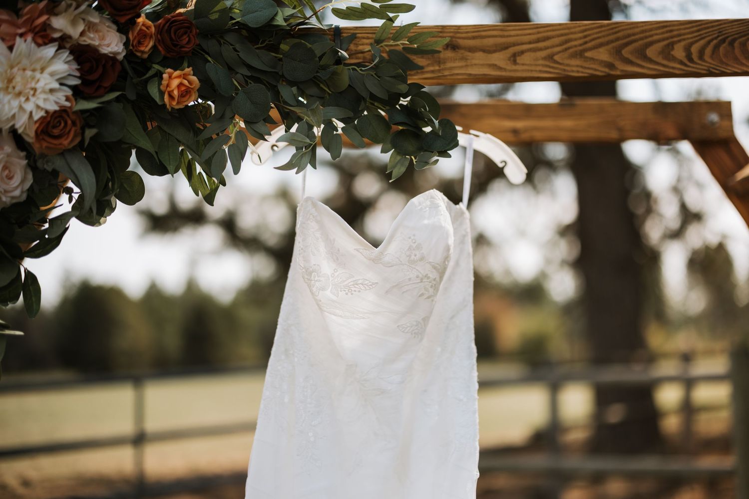 A delicate white wedding dress hangs from a wooden beam under greenery and flowers.