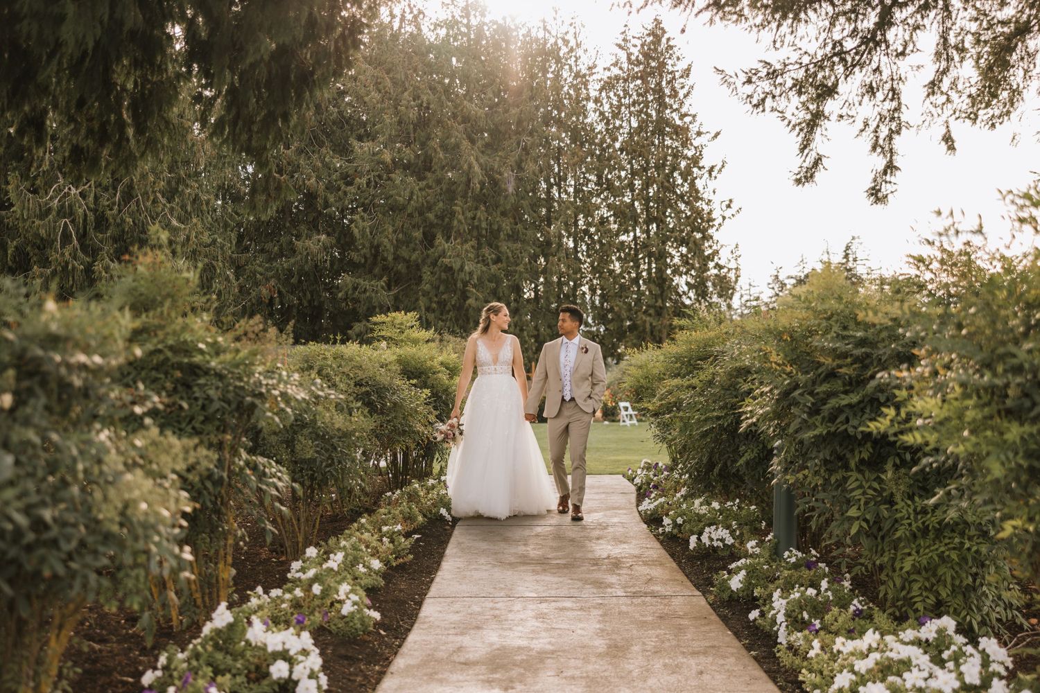 A couple walks down a garden path lined with white flowers and greenery.