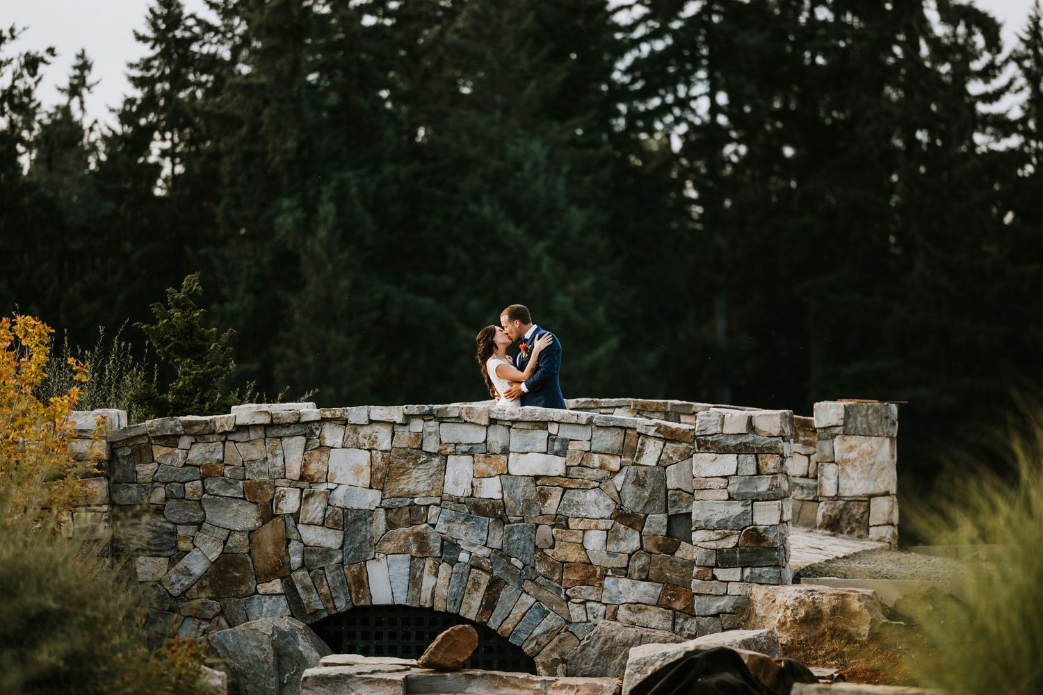 A romantic moment shared between newlyweds on a stone bridge with evergreen forest backdrop.