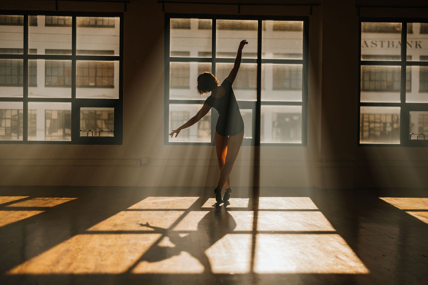 Silhouette of dancer in sunlit studio with large windows casting dramatic shadows.