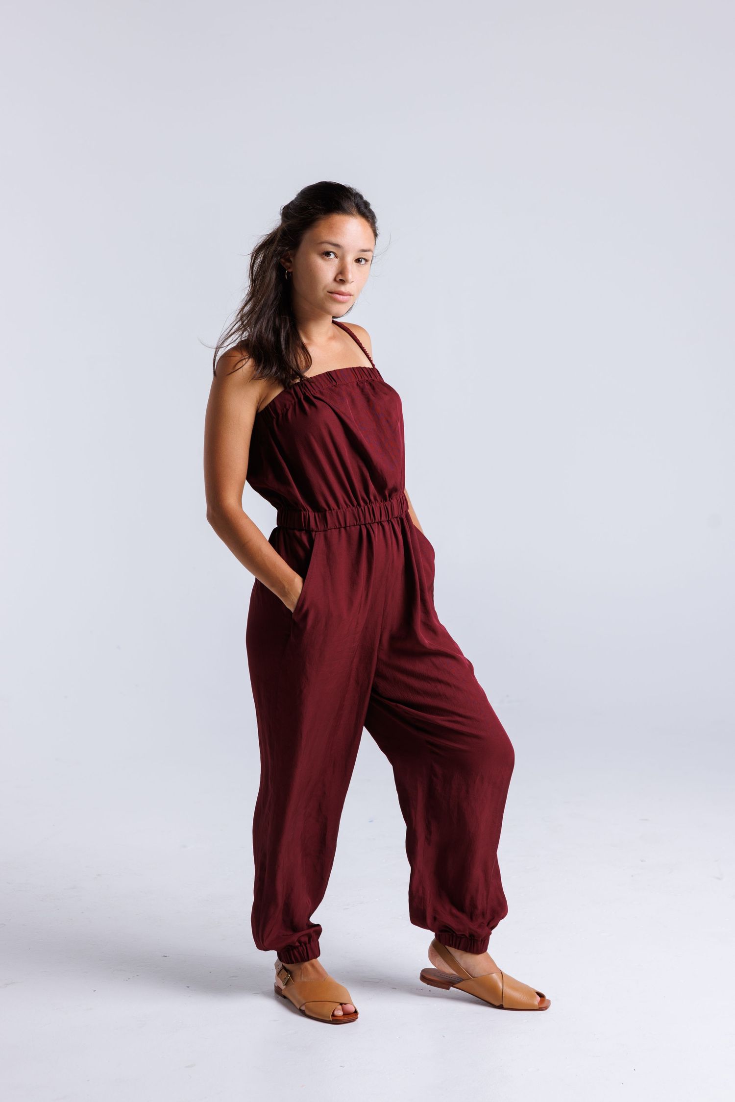 Model wearing burgundy jumpsuit poses against white background.
