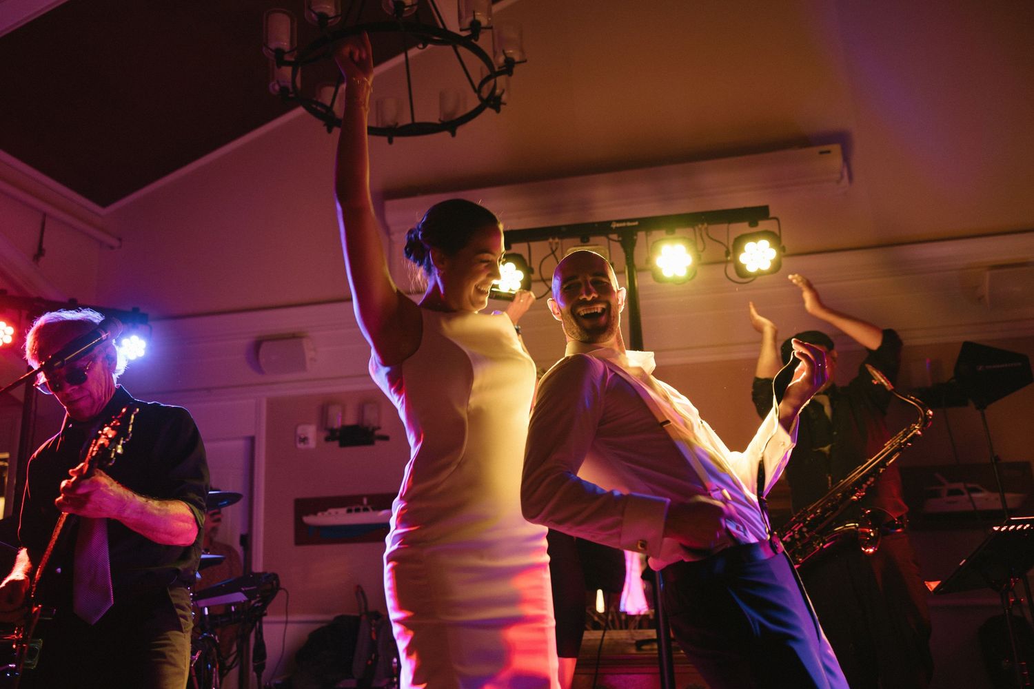 Bride and groom dancing joyfully on stage with live band musicians playing saxophone at a wedding reception.
