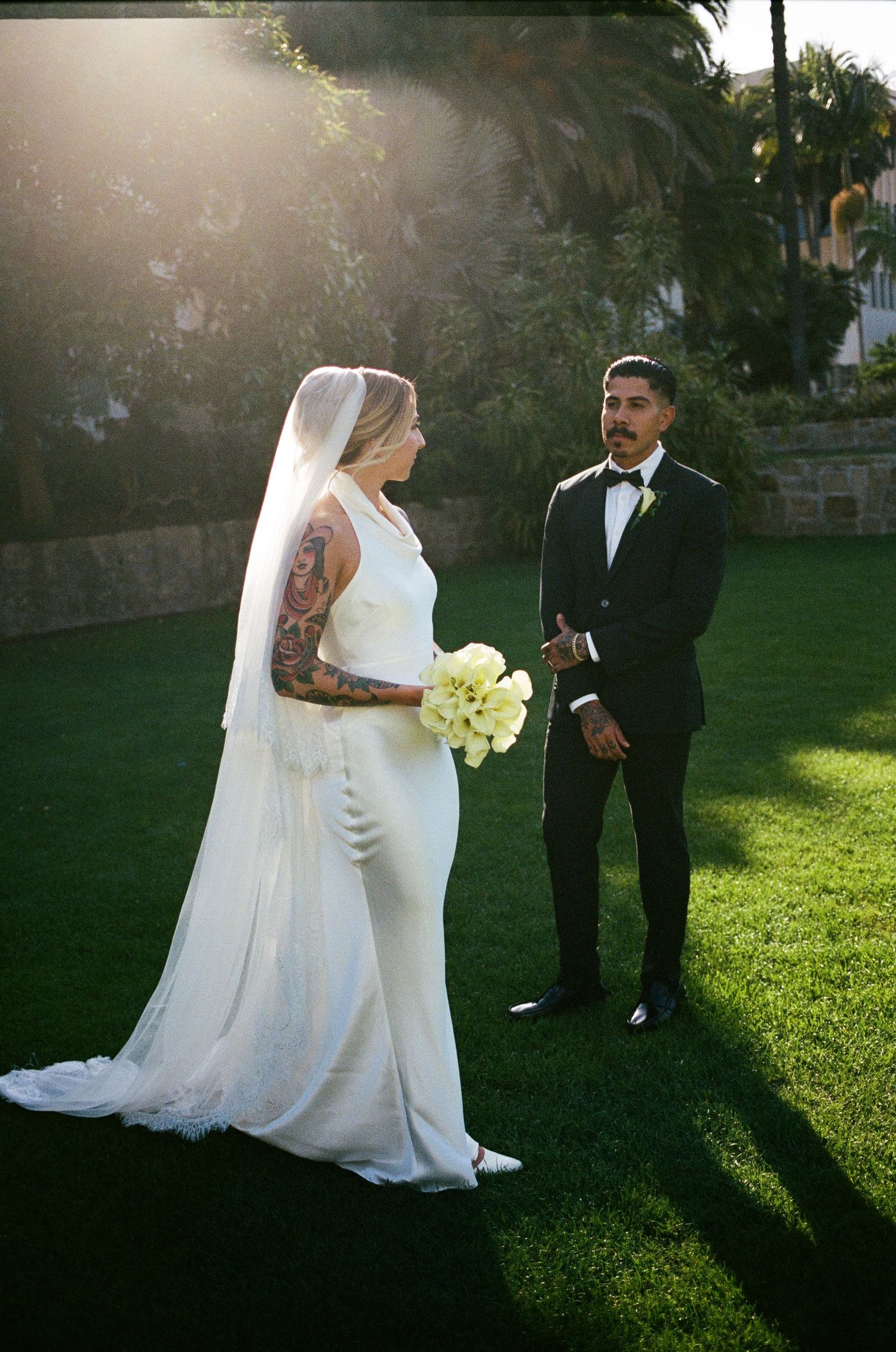 Bride in white gown with veil and tattoos holds bouquet facing groom in black tuxedo on green lawn at sunset.