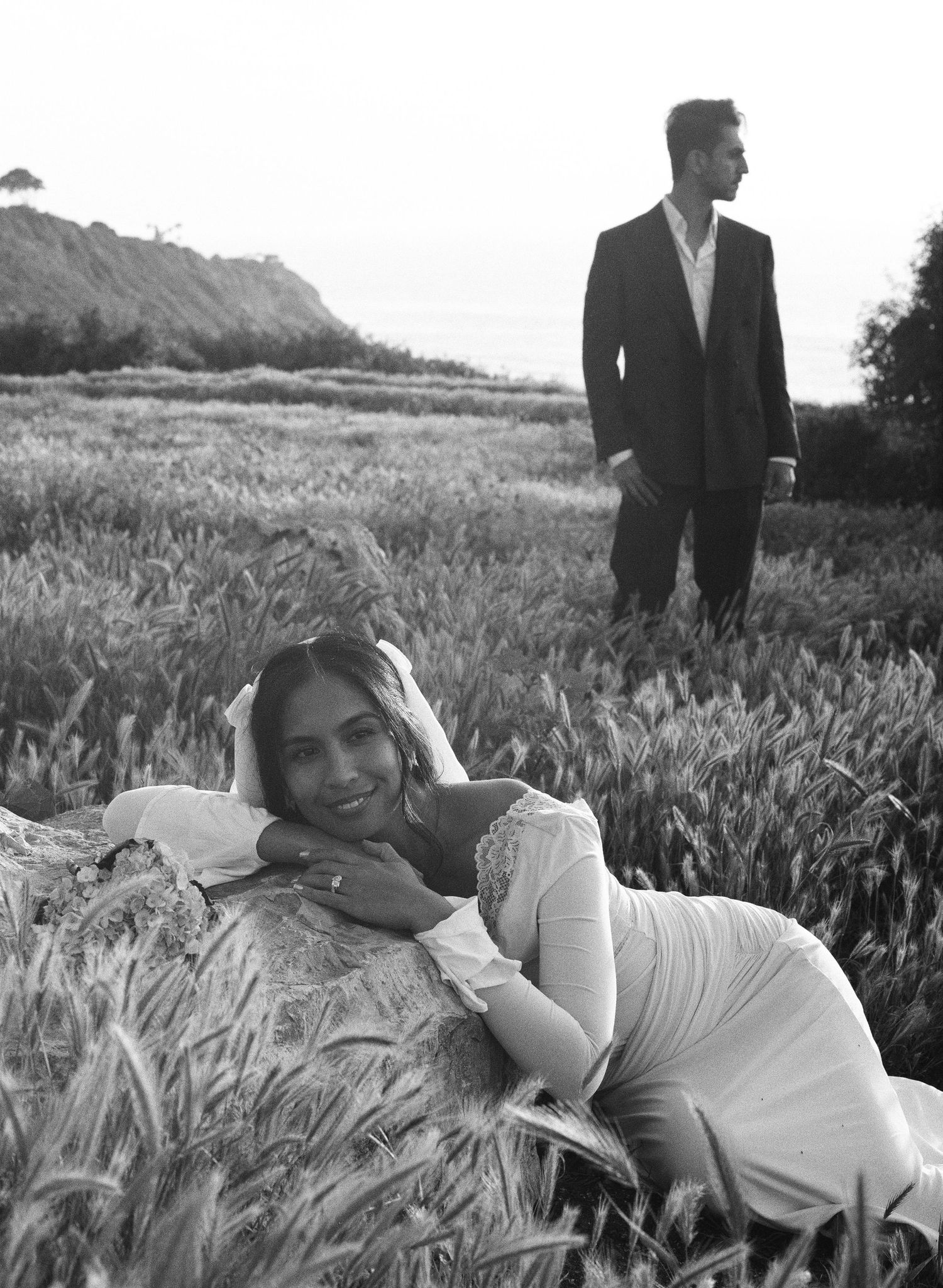 Black and white photo of a woman in white dress lying in wheat field, man in suit standing behind her.