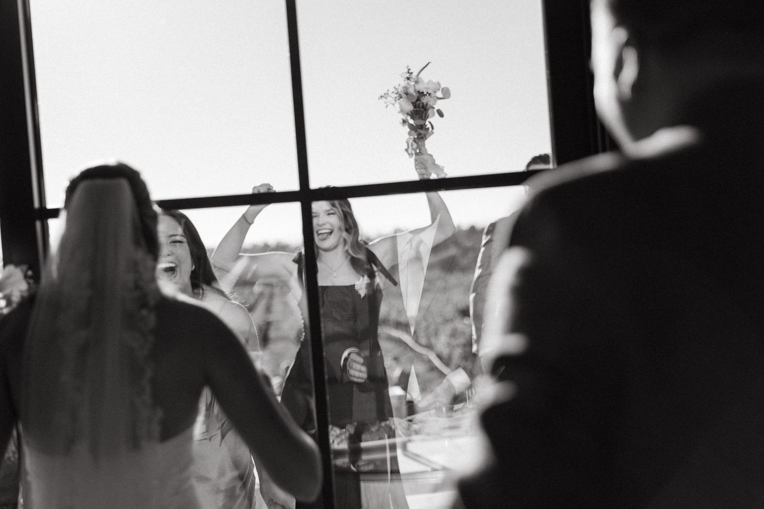 Black and white wedding photo of bride and bridesmaids celebrating joyfully, raising arms with bouquet near large windows.