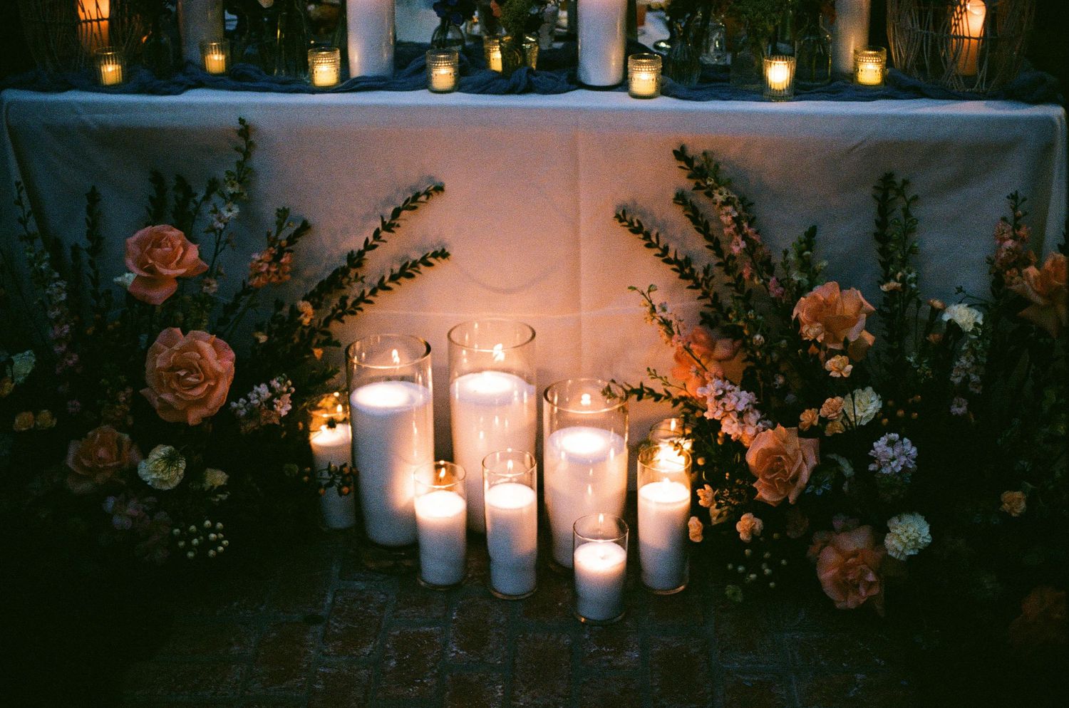 Romantic wedding table with white pillar candles, blush roses, and lush greenery arrangements in warm candlelight.