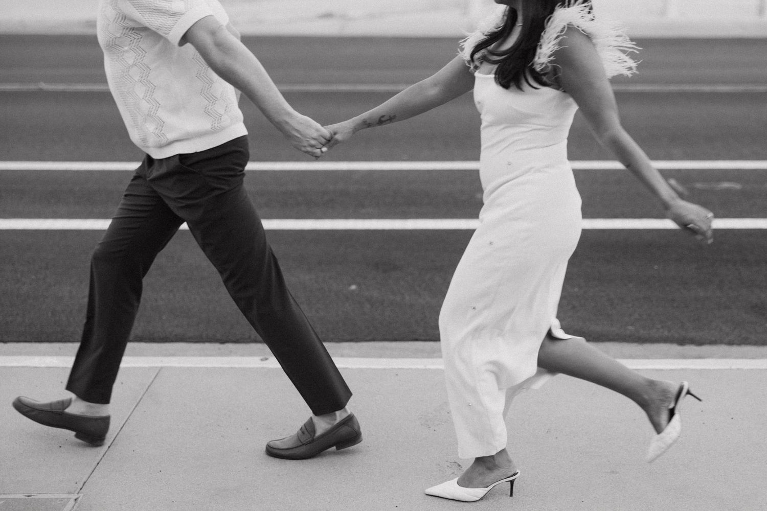 Black and white photo of a couple holding hands and walking together, dressed in wedding attire on a paved surface.
