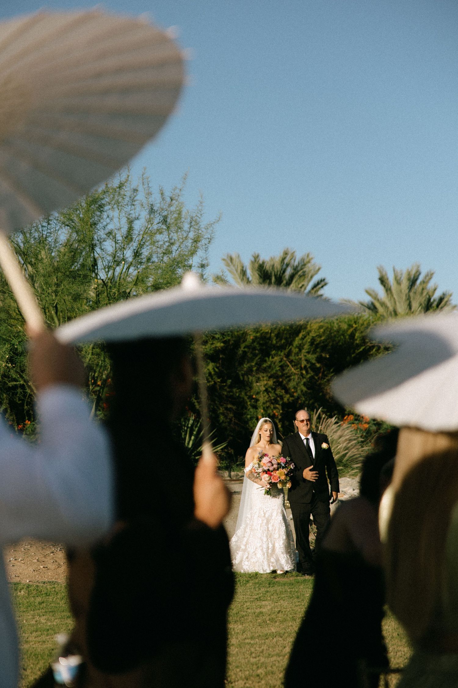 Bride walks down outdoor aisle with escort as guests hold white parasols at sunny garden wedding ceremony.
