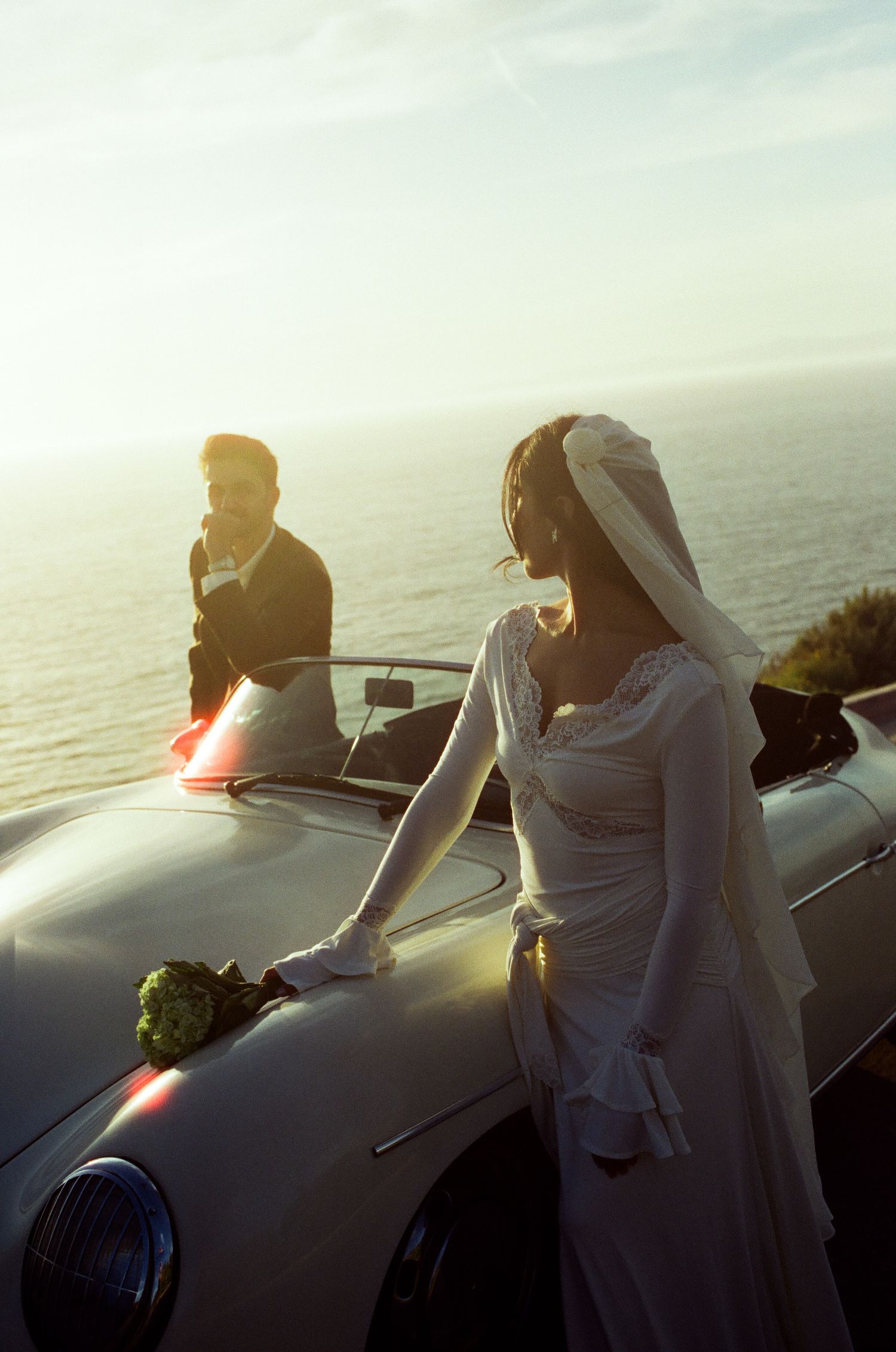 Bride in white lace gown and veil poses by vintage convertible car with groom, ocean horizon backdrop at sunset.