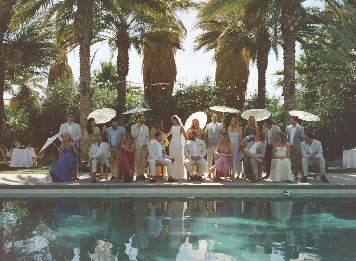 Wedding party posing by a pool surrounded by palm trees, guests in white attire holding parasols at an outdoor ceremony.