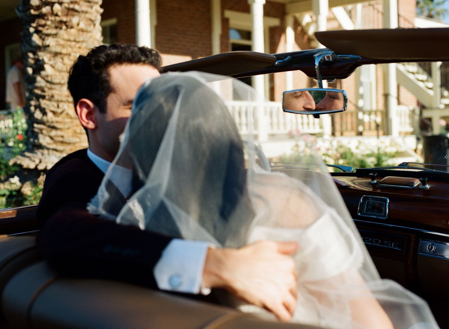 Newlywed couple sharing a romantic kiss in a vintage car, bride in white veil, groom in suit on wedding day.