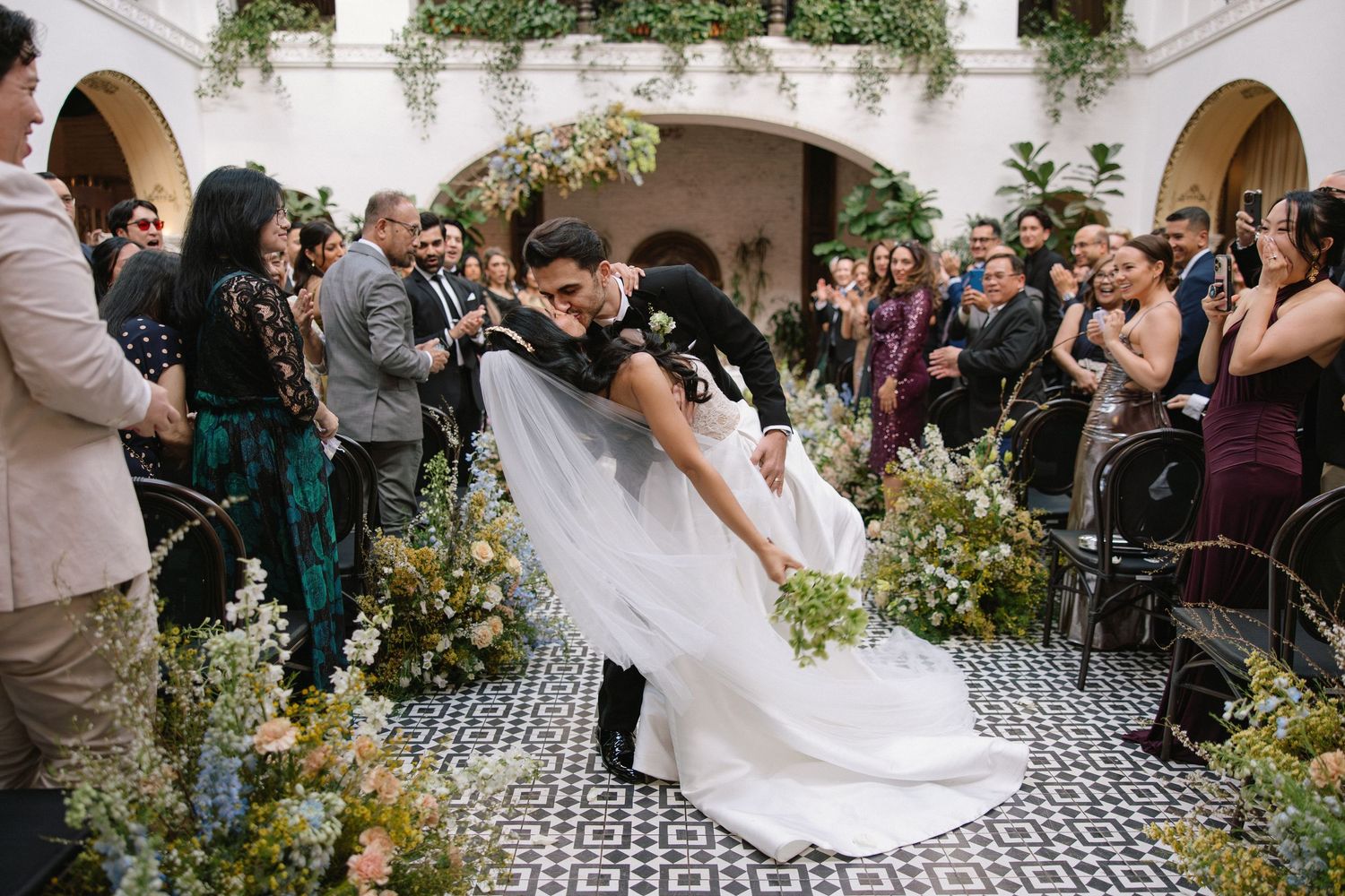 Bride and groom share romantic dip kiss at outdoor wedding ceremony surrounded by lush floral arrangements and guests.