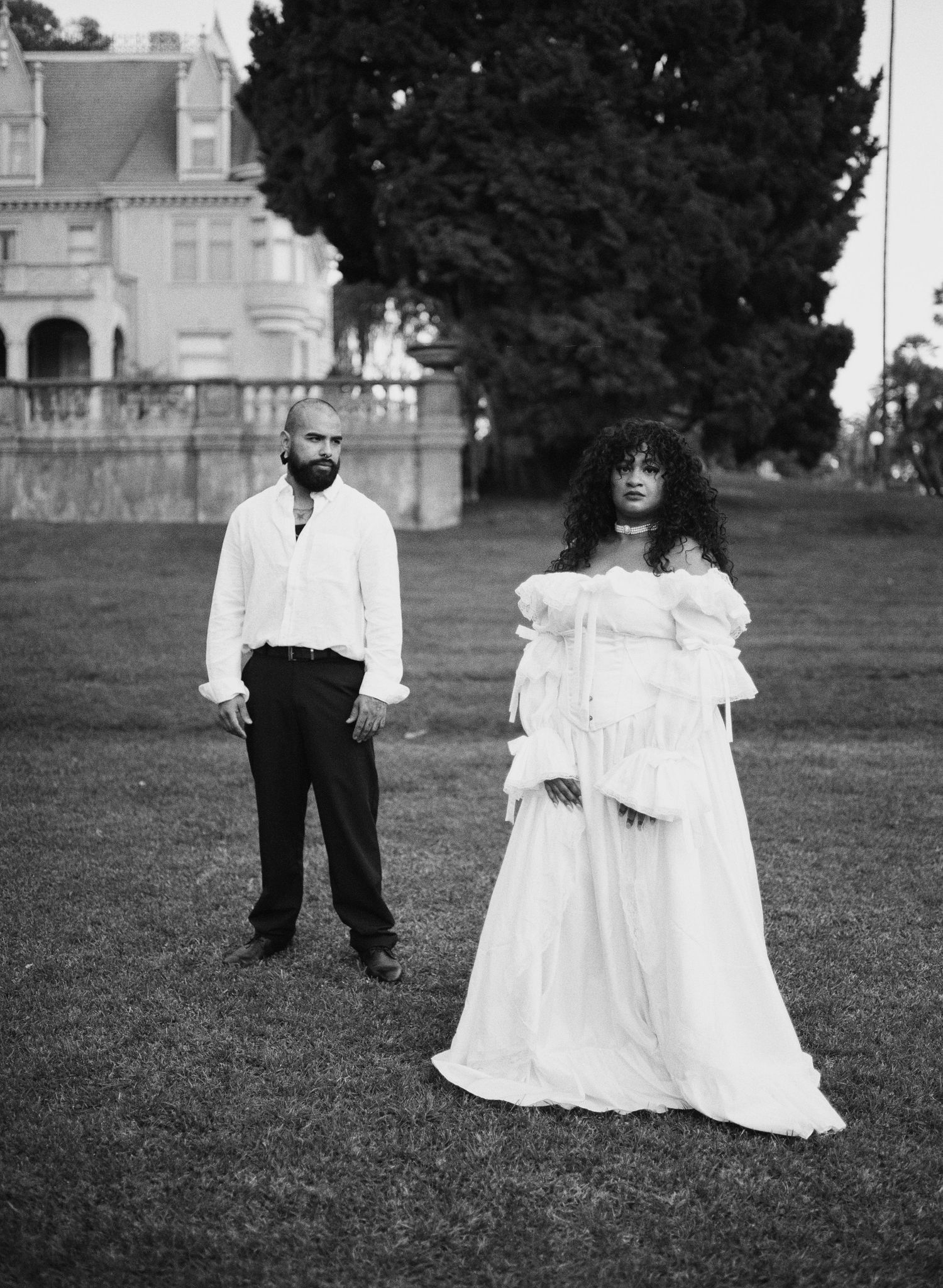 Black and white photo of a couple in white attire standing on a lawn before a grand historic mansion.