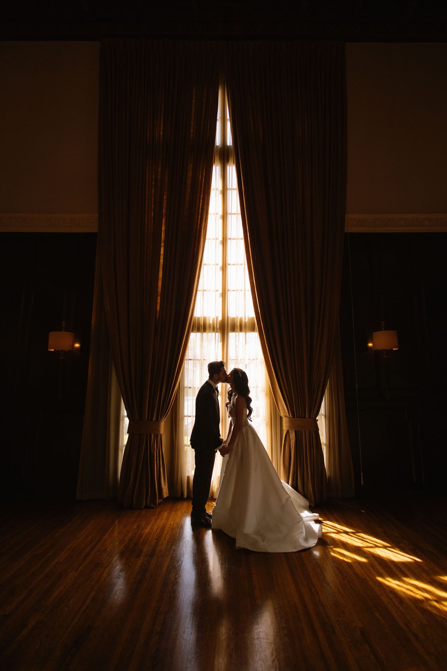 Bride and groom silhouetted kissing before grand curtained window with golden backlight on hardwood floor.