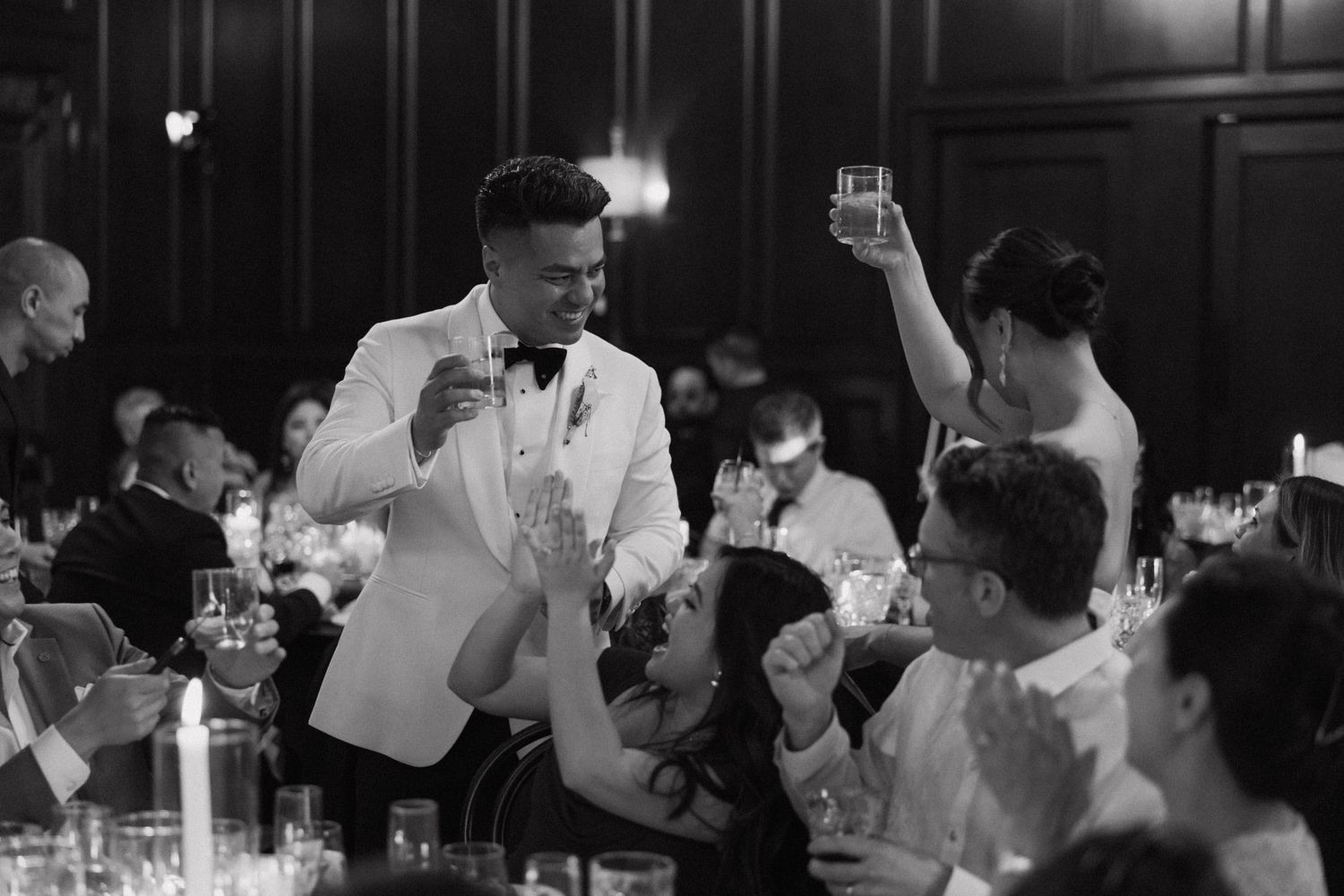 Groom in white tuxedo raises glass during elegant candlelit wedding reception toast in black and white photo.