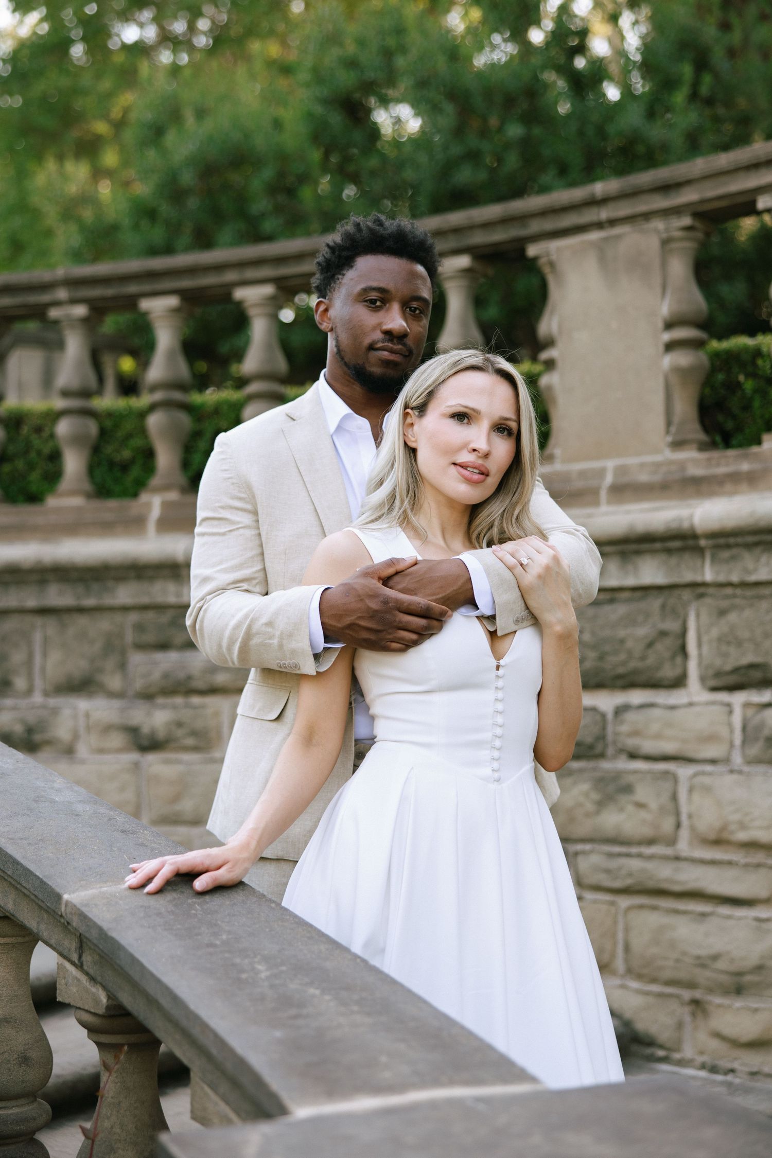 Interracial couple poses elegantly at stone balustrade, groom in beige suit embracing bride in white dress outdoors.