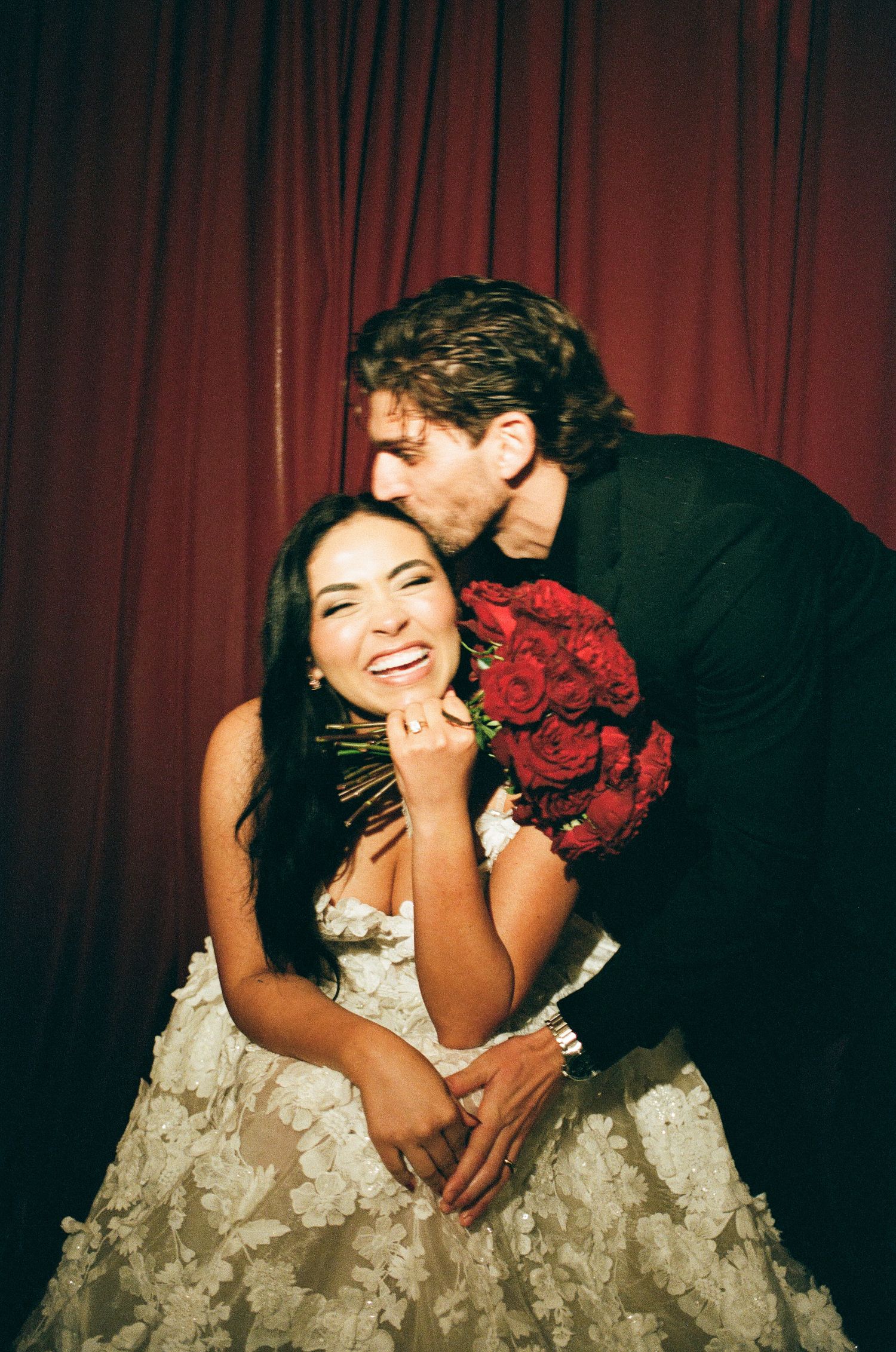 A joyful bride in a floral gown holds red roses as a groom in black kisses her cheek against a red curtain backdrop.