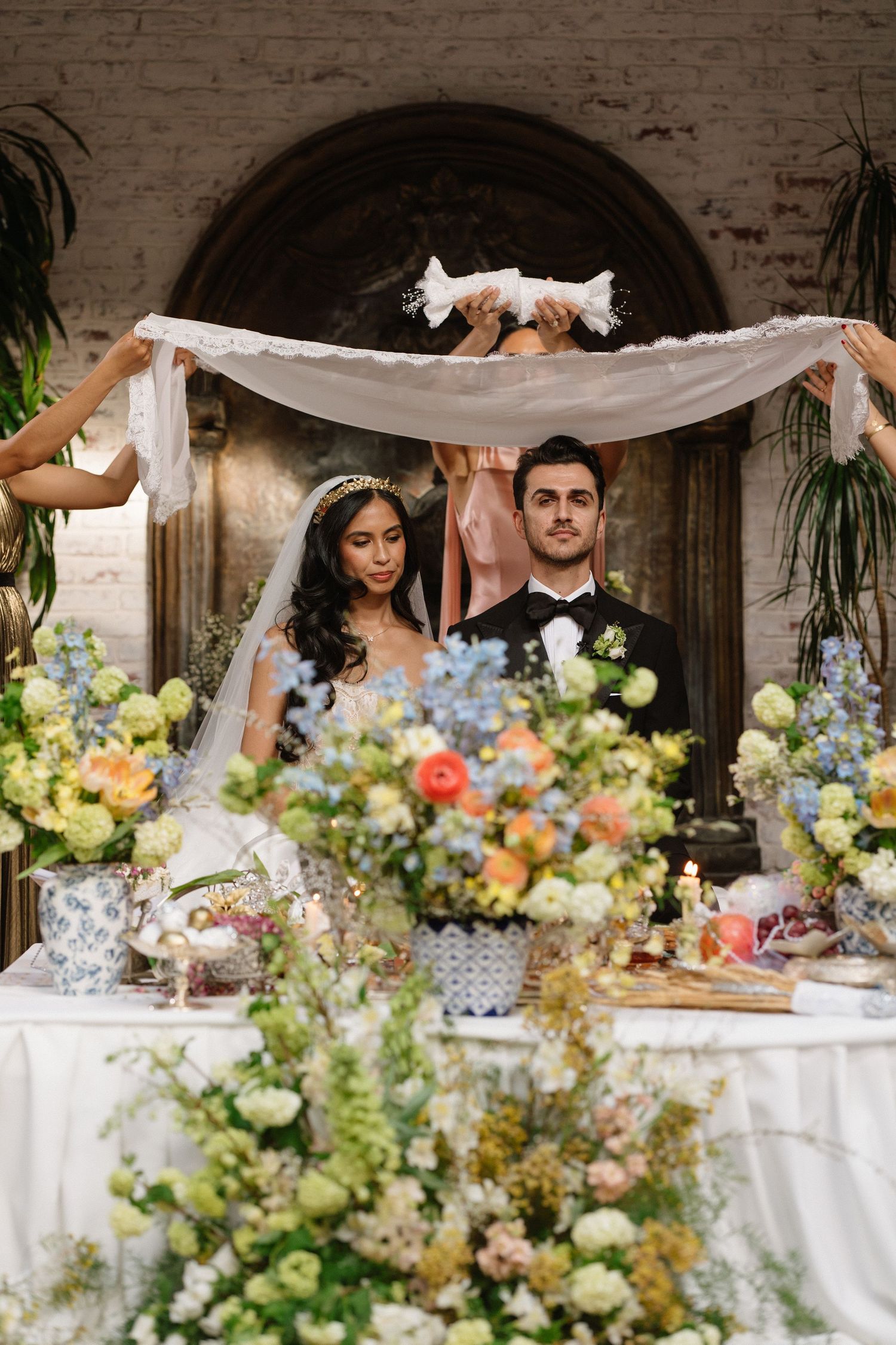 Bride and groom stand beneath a white fabric canopy surrounded by lush floral arrangements during an elegant wedding ceremony.