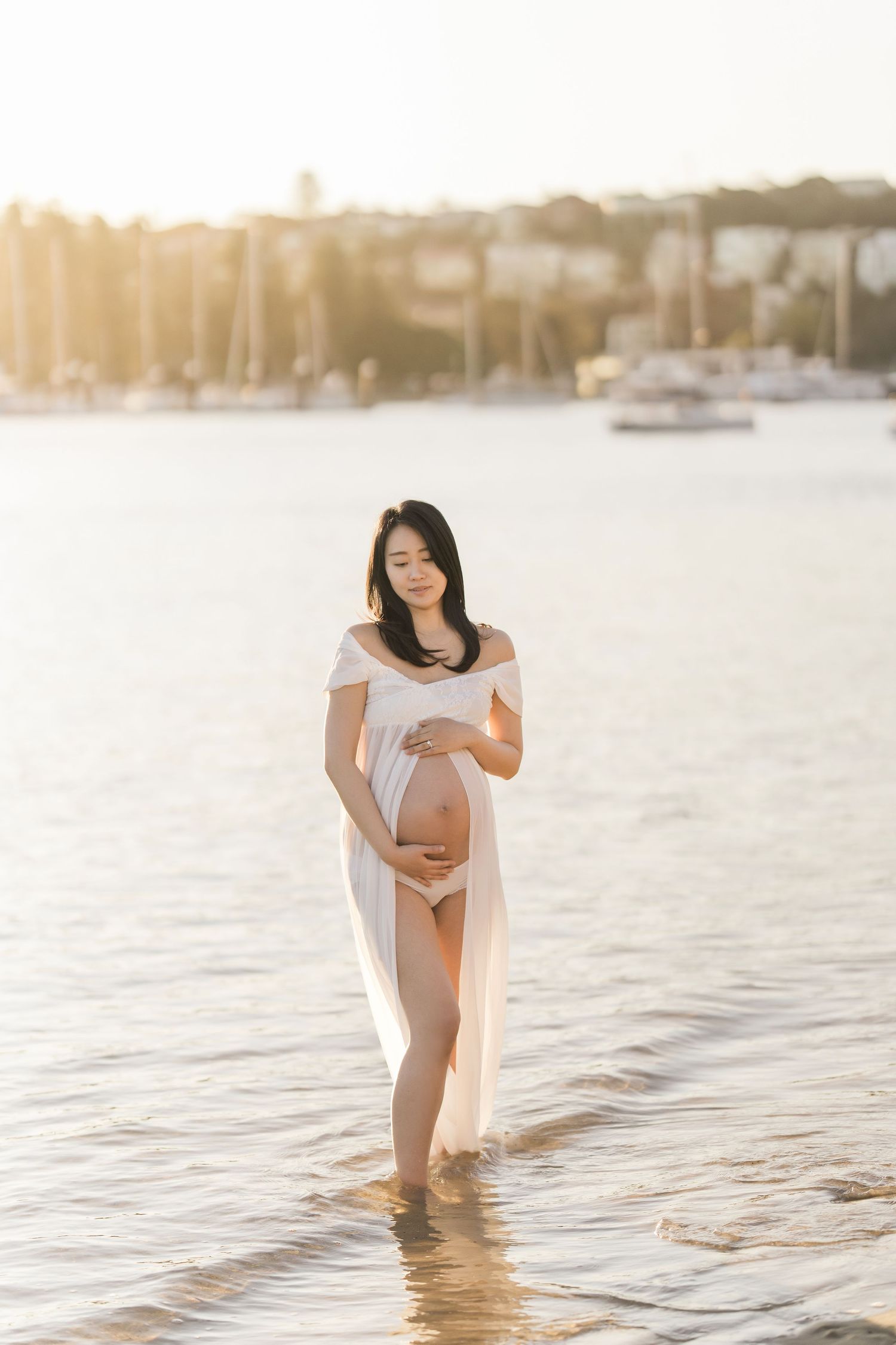 Pregnant woman in white sheer dress standing in shallow water at golden hour, cradling her baby bump near a marina.