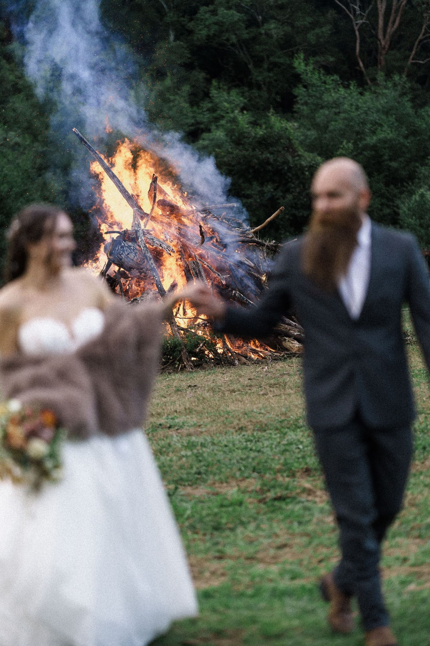 Bride and groom stand before a large bonfire at an outdoor rustic wedding ceremony surrounded by lush green trees.