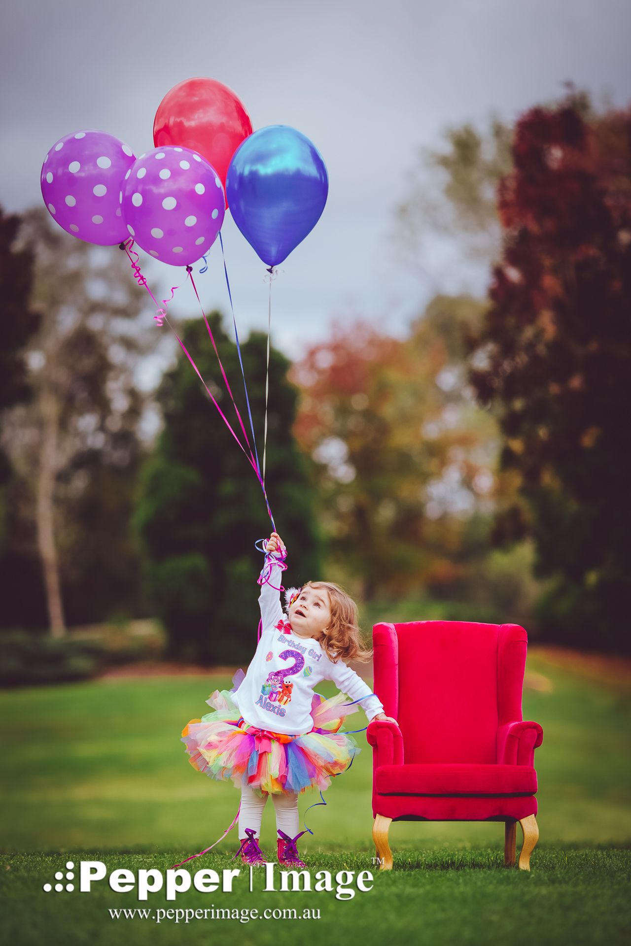 A toddler girl in a colorful tutu holds balloons near a red chair outdoors on her 2nd birthday.