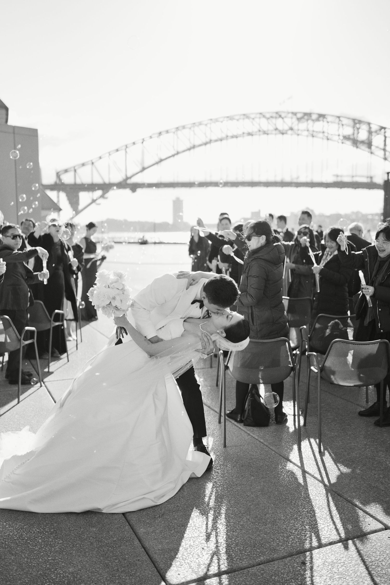 Bride and groom share a romantic dip kiss at outdoor wedding ceremony with Sydney Harbour Bridge in background.