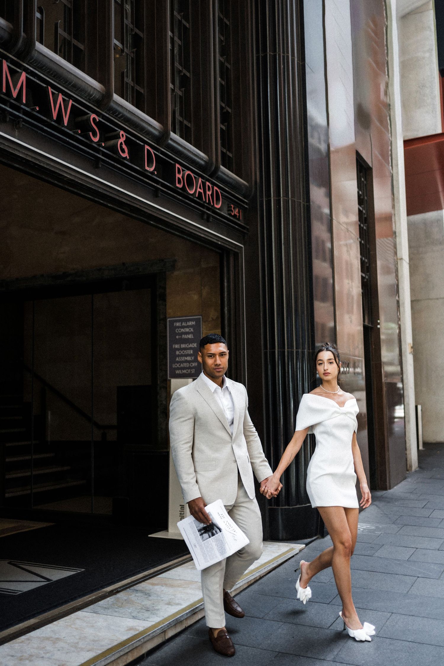Elegant couple in white mini dress and beige suit holding hands outside a grand city building.