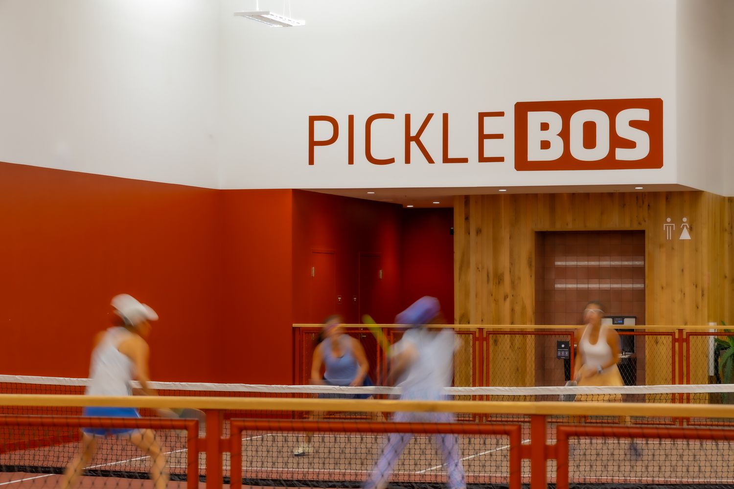 Players enjoy pickleball at PickleBos indoor court facility with vibrant red and white branding on the wall.