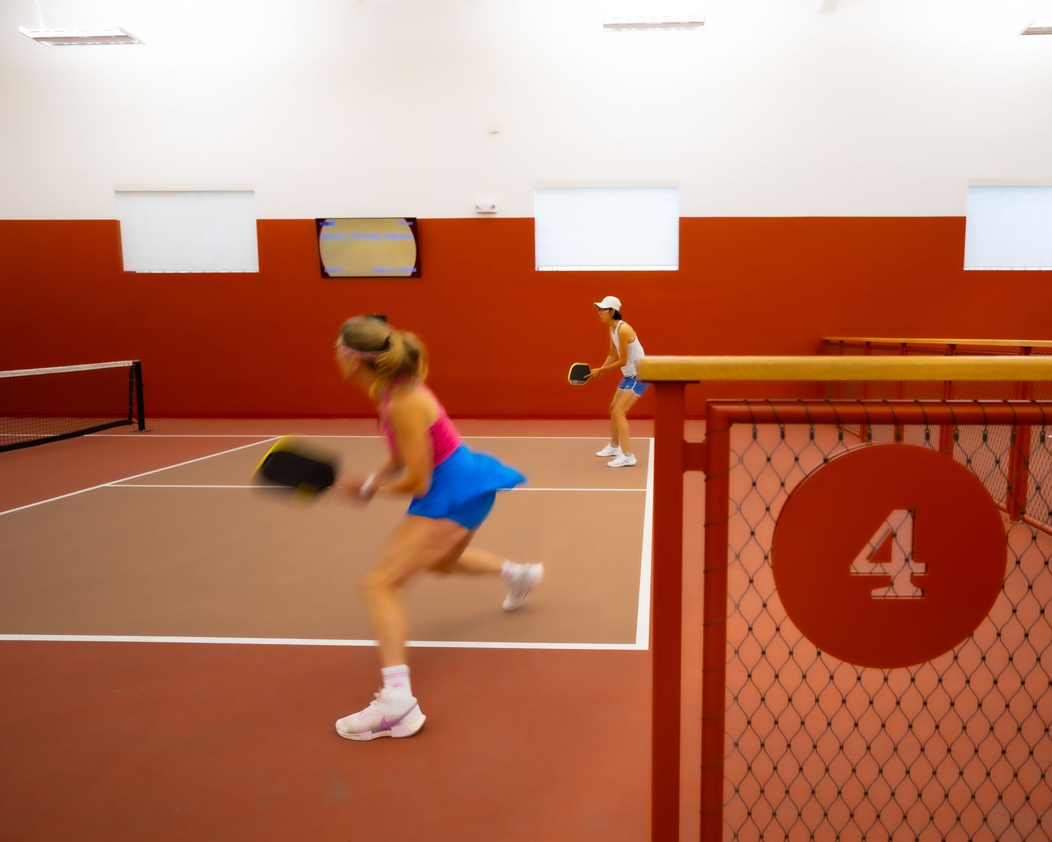 Two women playing indoor platform tennis on a red court, with a number 4 sign on the chain-link fence divider.