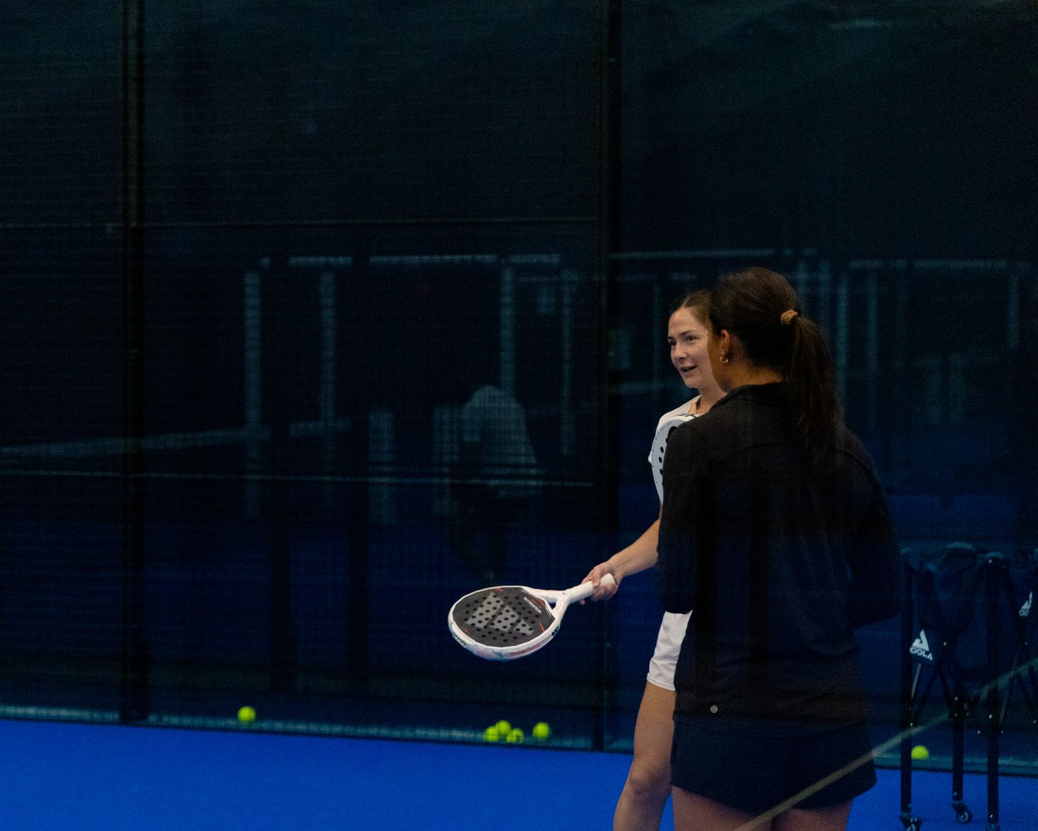 Woman holding a padel racket on a blue indoor court with tennis balls on the ground.