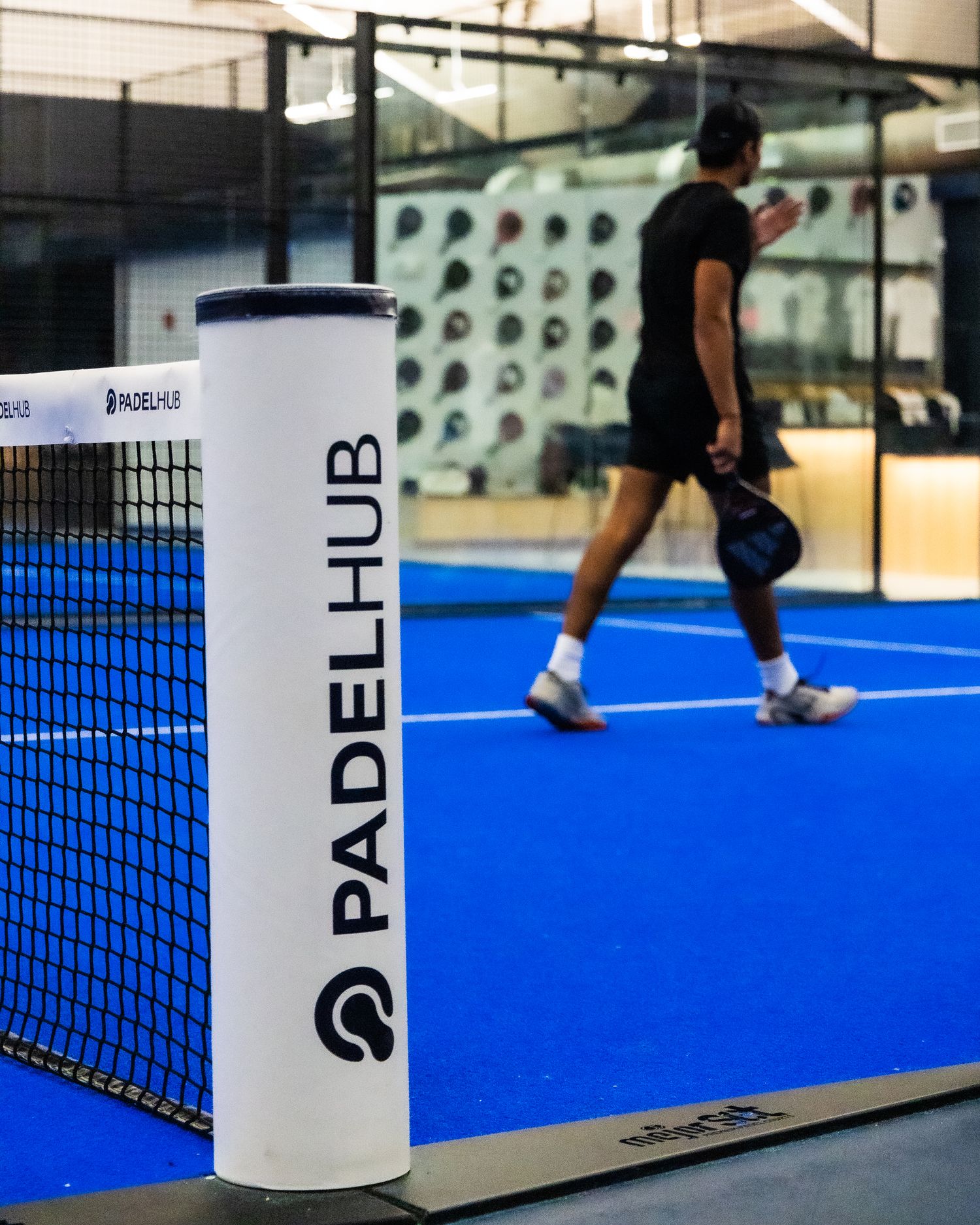 Padel Hub branded net post on a blue indoor padel court with a player walking in background.