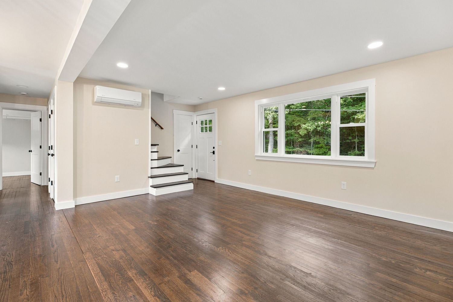 Bright open living room with dark hardwood floors, white walls, recessed lighting, staircase, and a large window with garden views.