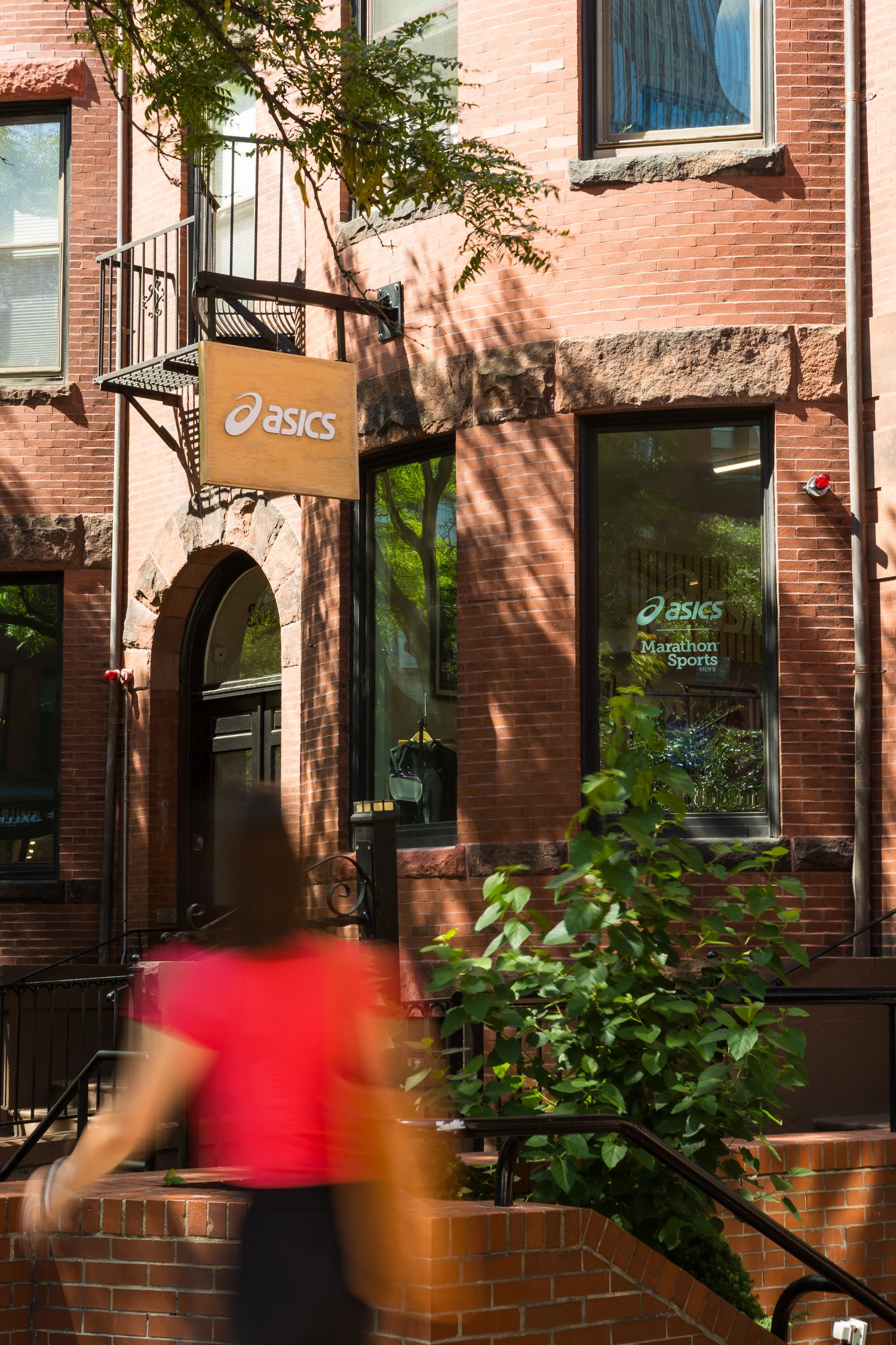 Asics Marathon Sports store in red brick Boston building with blurred pedestrian in red shirt.