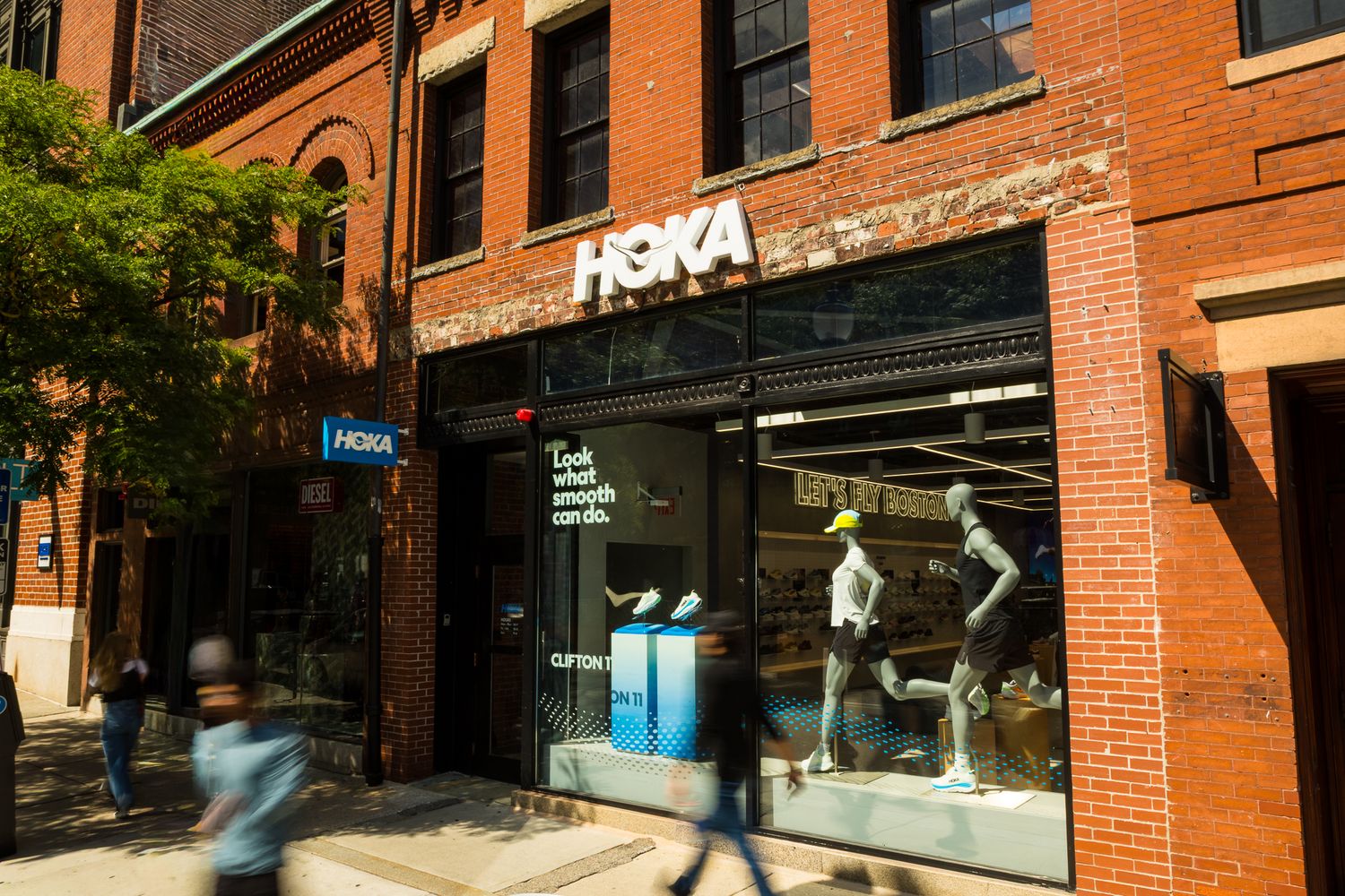 HOKA shoe store exterior in red brick building with mannequins and shoppers passing by.