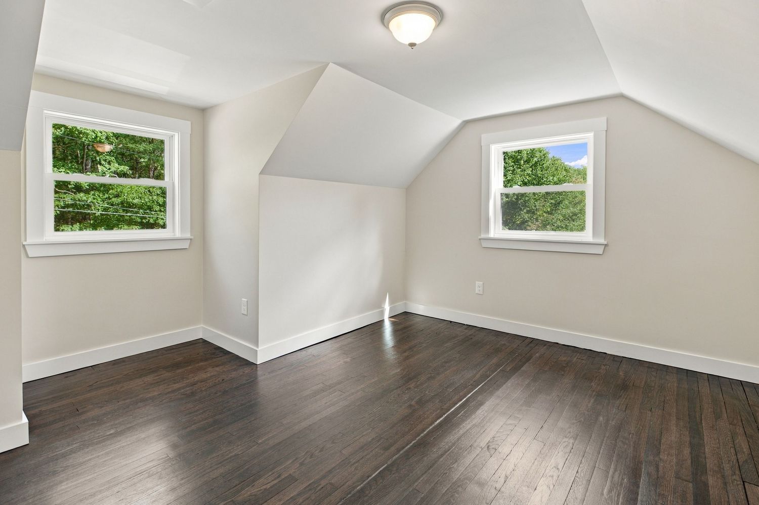 Bright attic bedroom with vaulted ceilings, dark hardwood floors, white trim, and two windows with green tree views.