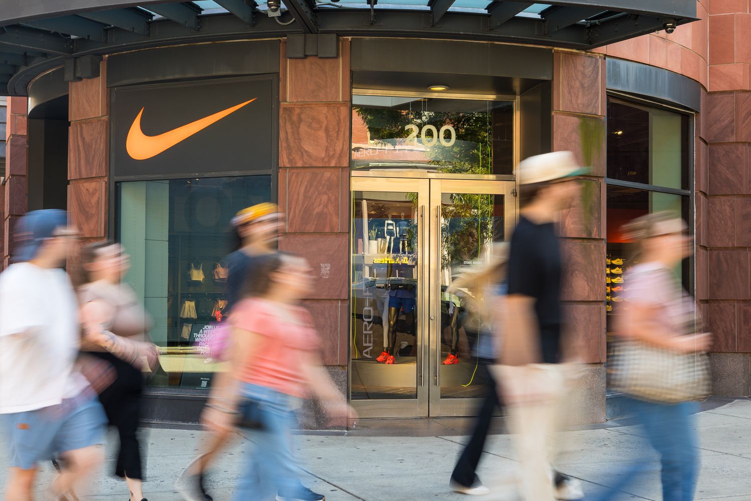 Busy Nike retail store exterior with orange swoosh logo and shoppers walking past in motion blur.