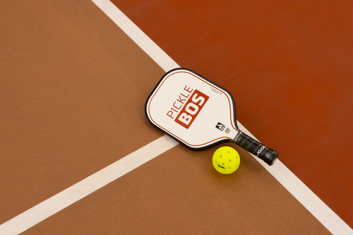 Pickleball paddle with PICKLE BDS branding beside a yellow ball on a brown court near white boundary lines.