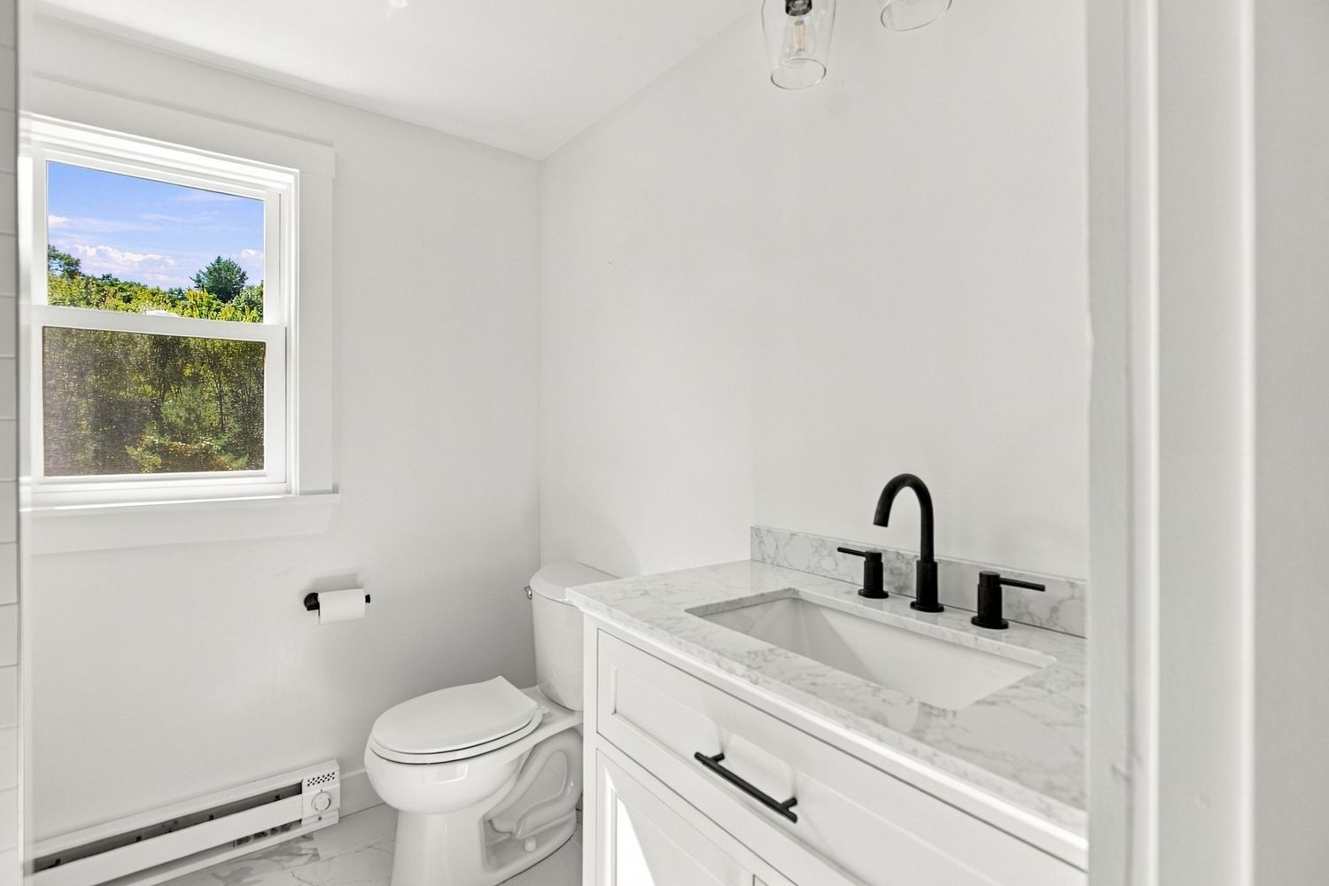 Bright white bathroom with marble vanity, black fixtures, toilet, baseboard heater, and a window with natural light.