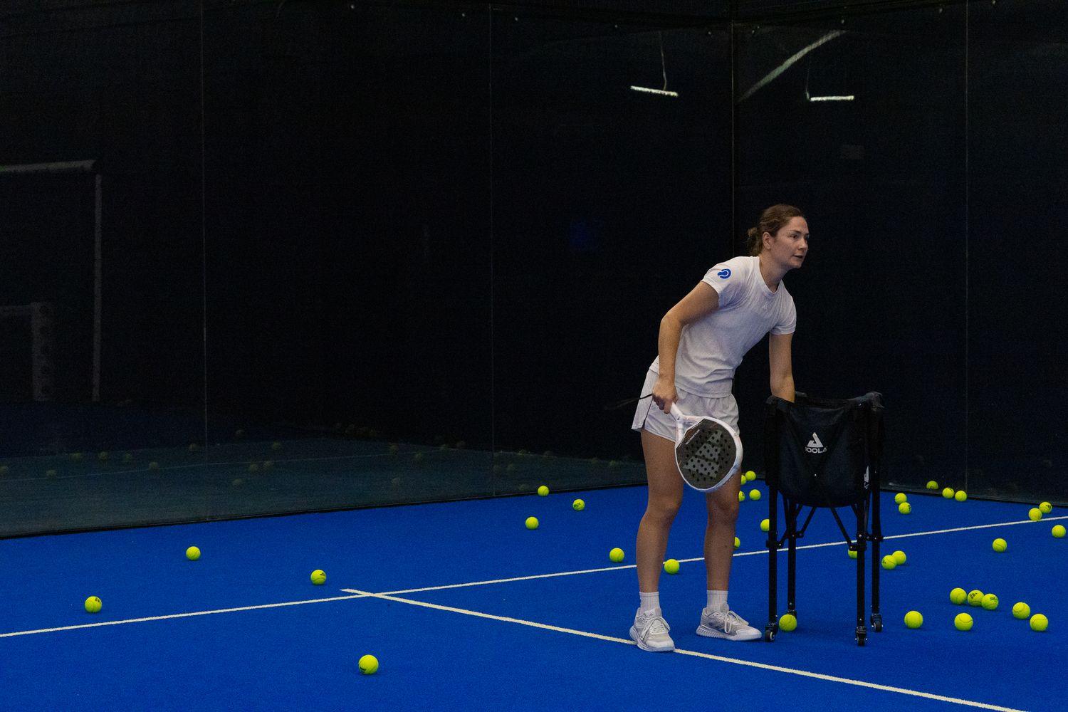 Female padel player ready to serve surrounded by yellow balls on a blue indoor court.