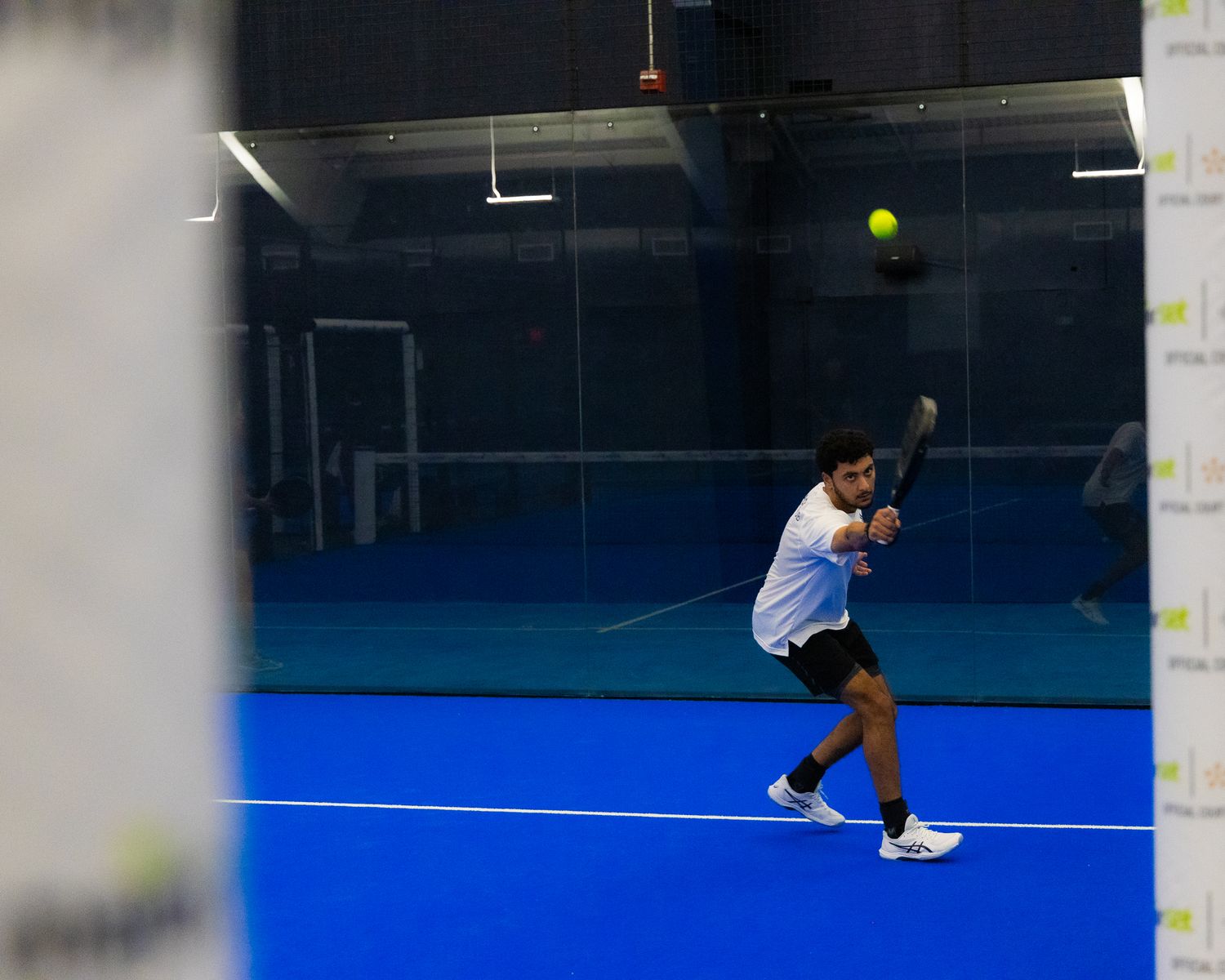 Padel tennis player executing a powerful shot on a blue indoor court through glass wall.