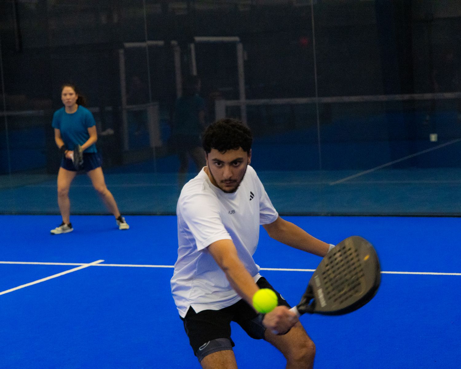 Male padel player hitting a backhand shot on a blue indoor court with female partner.